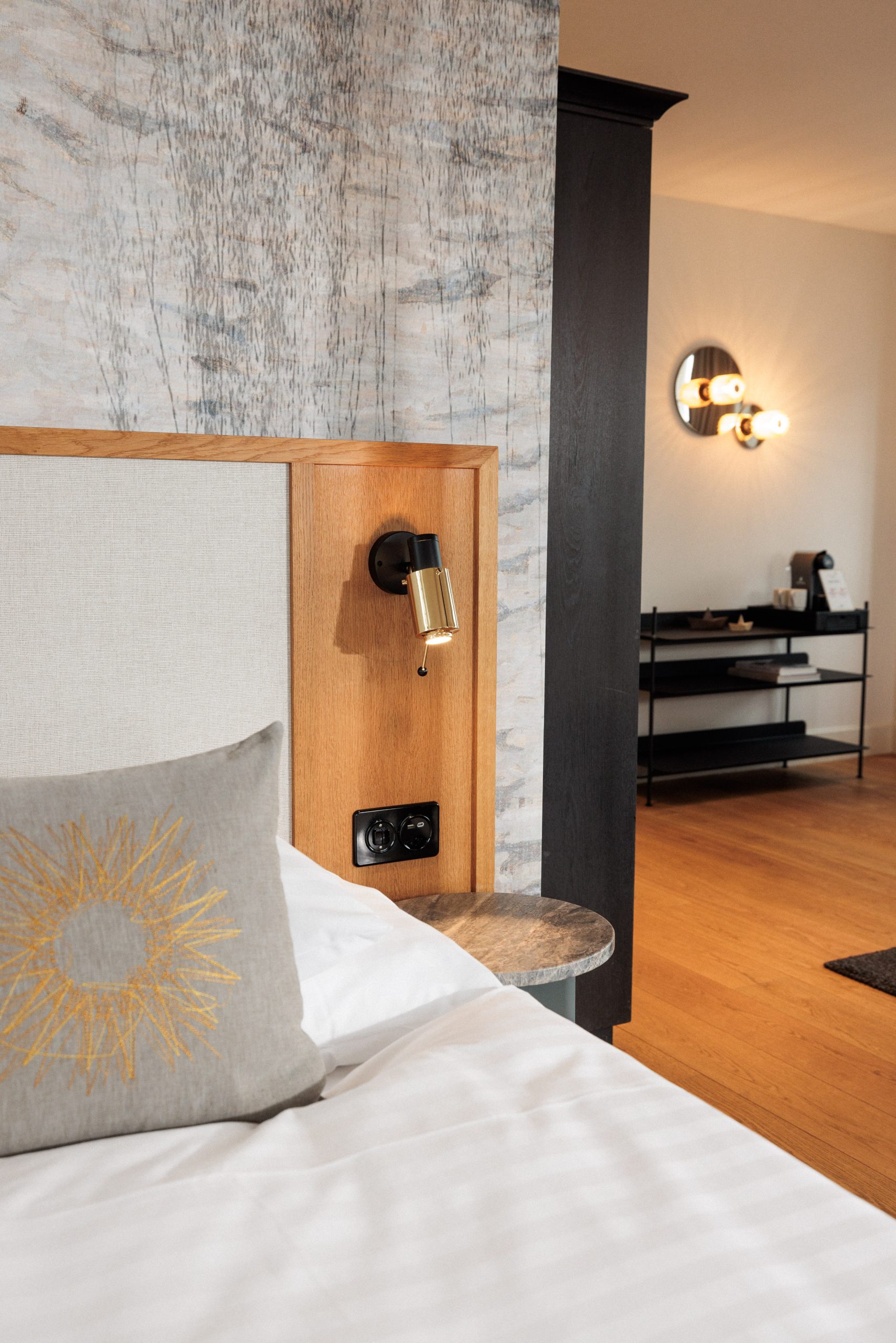 Ein modernes Boutiquehotelzimmer mit einer Nahaufnahme eines ordentlich gemachten Bettes, einem grauen Kissen mit gelbem Sonnenschliffmuster, einem hölzernen Kopfteil mit Leselicht und einem Blick auf stilvolle Wände und Regale im Hintergrund.