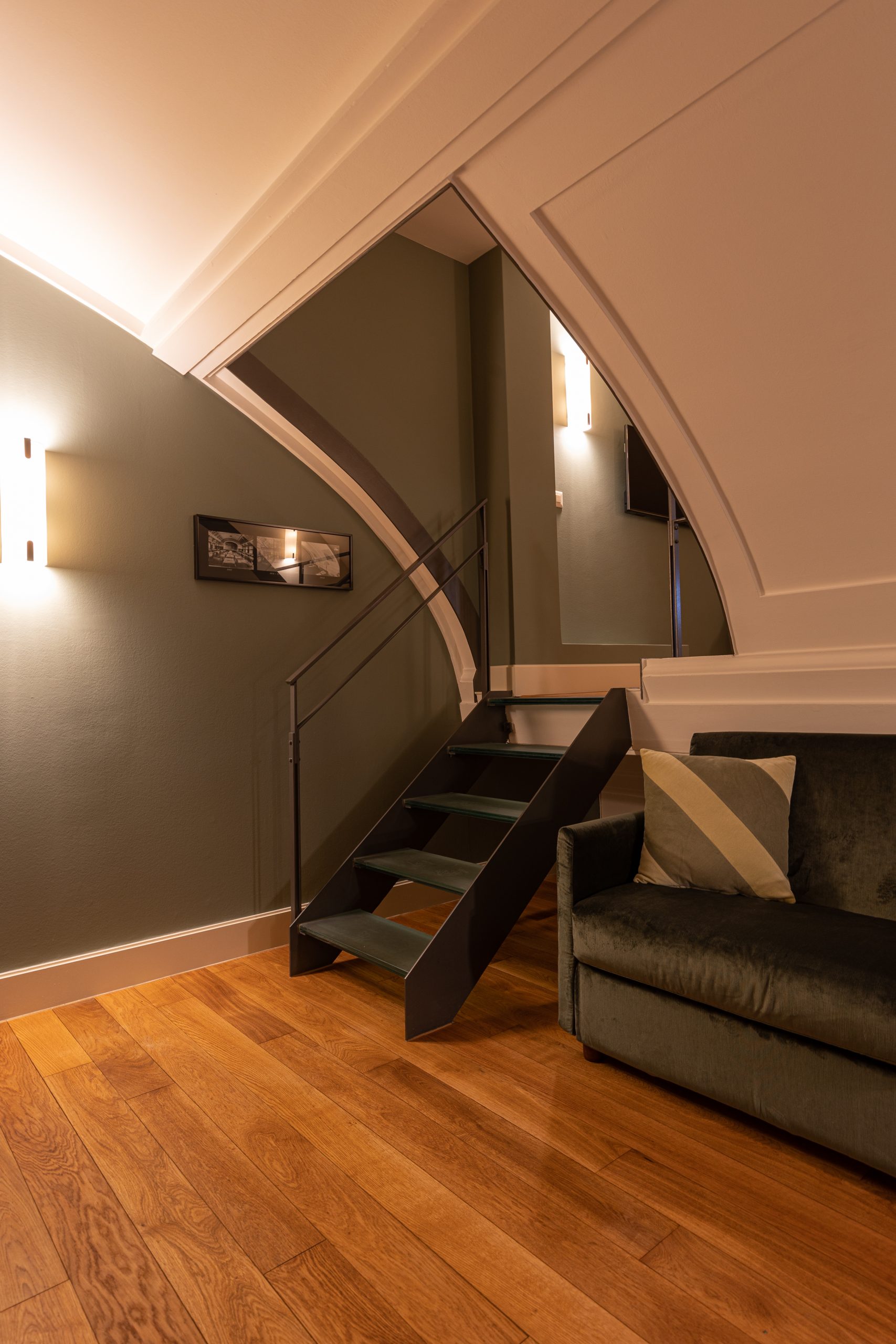 Ein moderner Wohnbereich in einem Boutiquehotel mit Holzfußboden, einem dunkelgrünen Samtsofa mit Kissen und einer eleganten schwarzen Metalltreppe. Sanfte Wandbeleuchtung und grüne Wände schaffen eine einladende, gemütliche Atmosphäre.