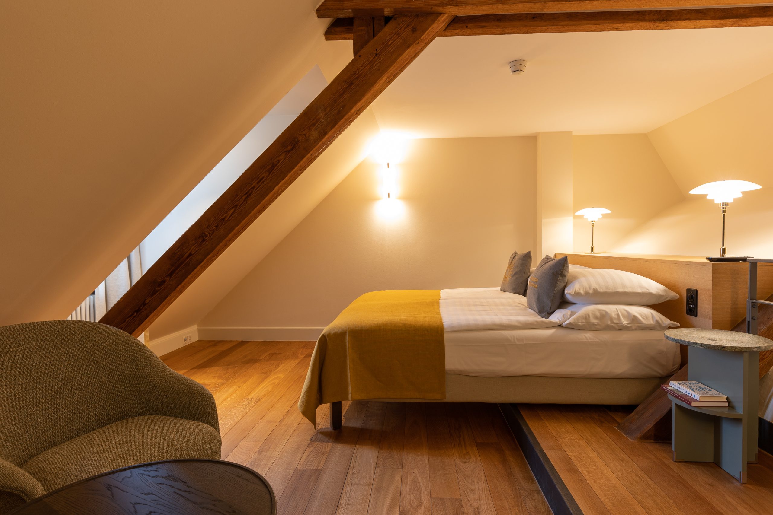 Ein gemütliches Zimmer im Dachgeschoss eines Boutiquehotels mit Holzbalken, einem Doppelbett mit weißer Bettwäsche und senffarbener Decke, sanfter Beleuchtung, einem kleinen runden Beistelltisch, einem Sessel und Parkettboden.