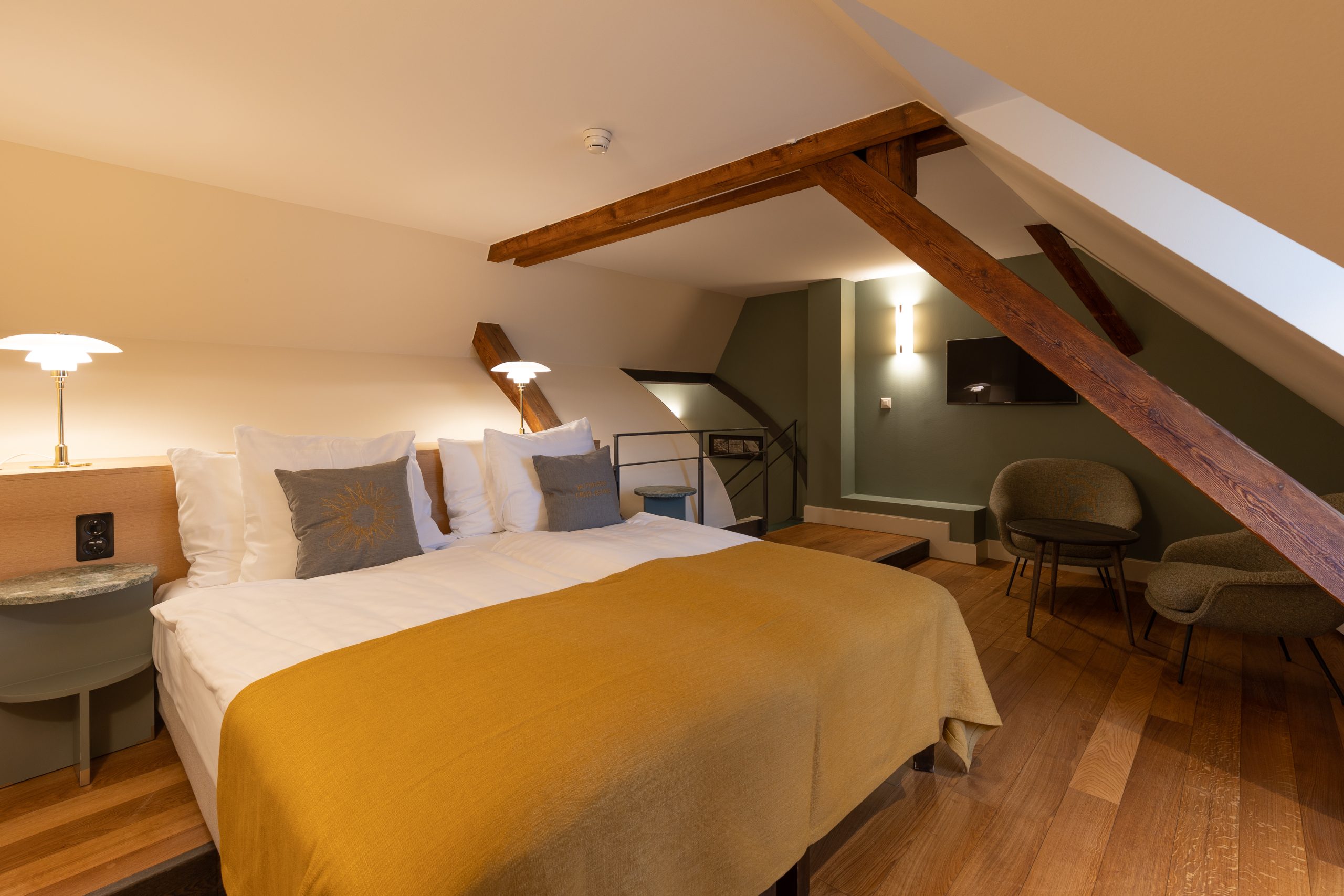 Ein gemütliches Boutiquehotelzimmer mit Doppelbett, weißer Bettwäsche, senfgelber Decke, Nachttischlampen, Holzbalken, schräger Decke, Sitzecke mit Stühlen und Tisch und warmer Beleuchtung.