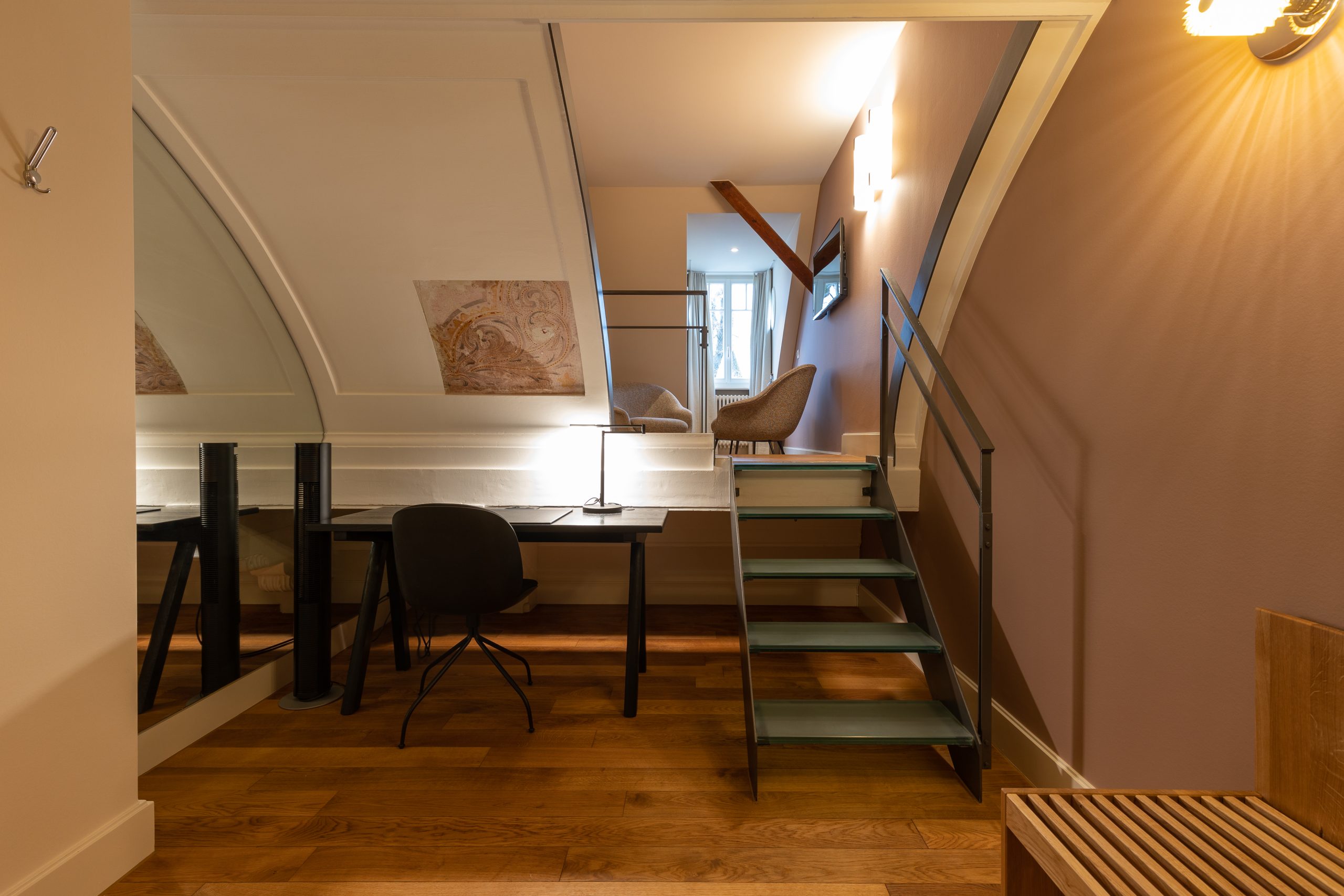 Ein kleines, modernes Zimmer in einem Boutiquehotel mit Holzfußboden, einem schwarzen Schreibtisch und Stuhl darunter und einer Metalltreppe, die zu einer gemütlichen Sitzecke an einem hellen Fenster führt. Sanfte Beleuchtung und neutrale Wände schaffen eine warme, einladende Atmosphäre.