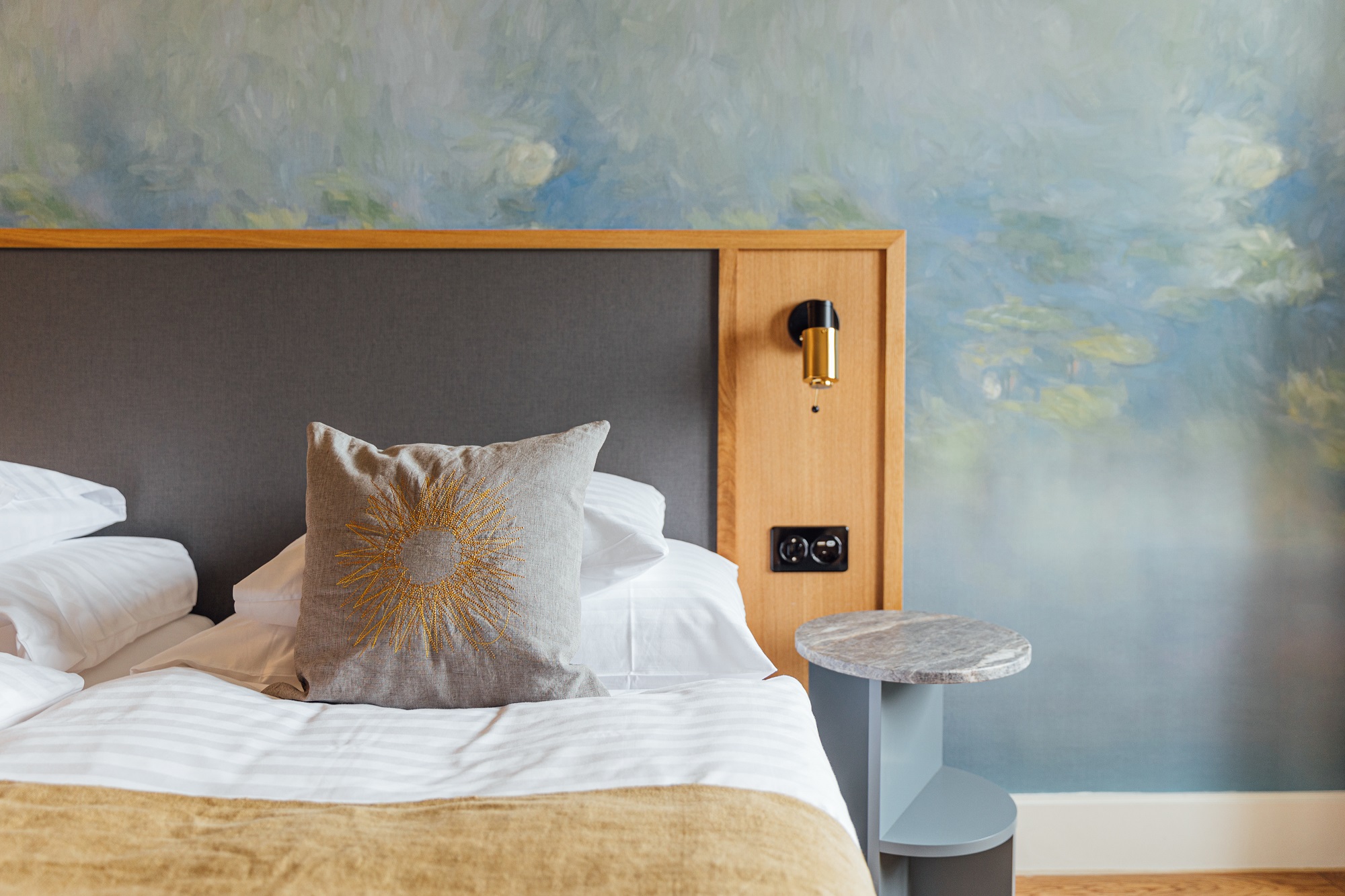 Ein ordentlich gemachtes Bett mit weißer Bettwäsche, einem grauen Kissen mit Sonnenschliff und einer hellbraunen Decke steht vor einem Kopfteil mit einer goldenen Wandlampe, neben einem kleinen runden Beistelltisch und einer blau getönten Kunstwand in diesem schicken Boutiquehotel.