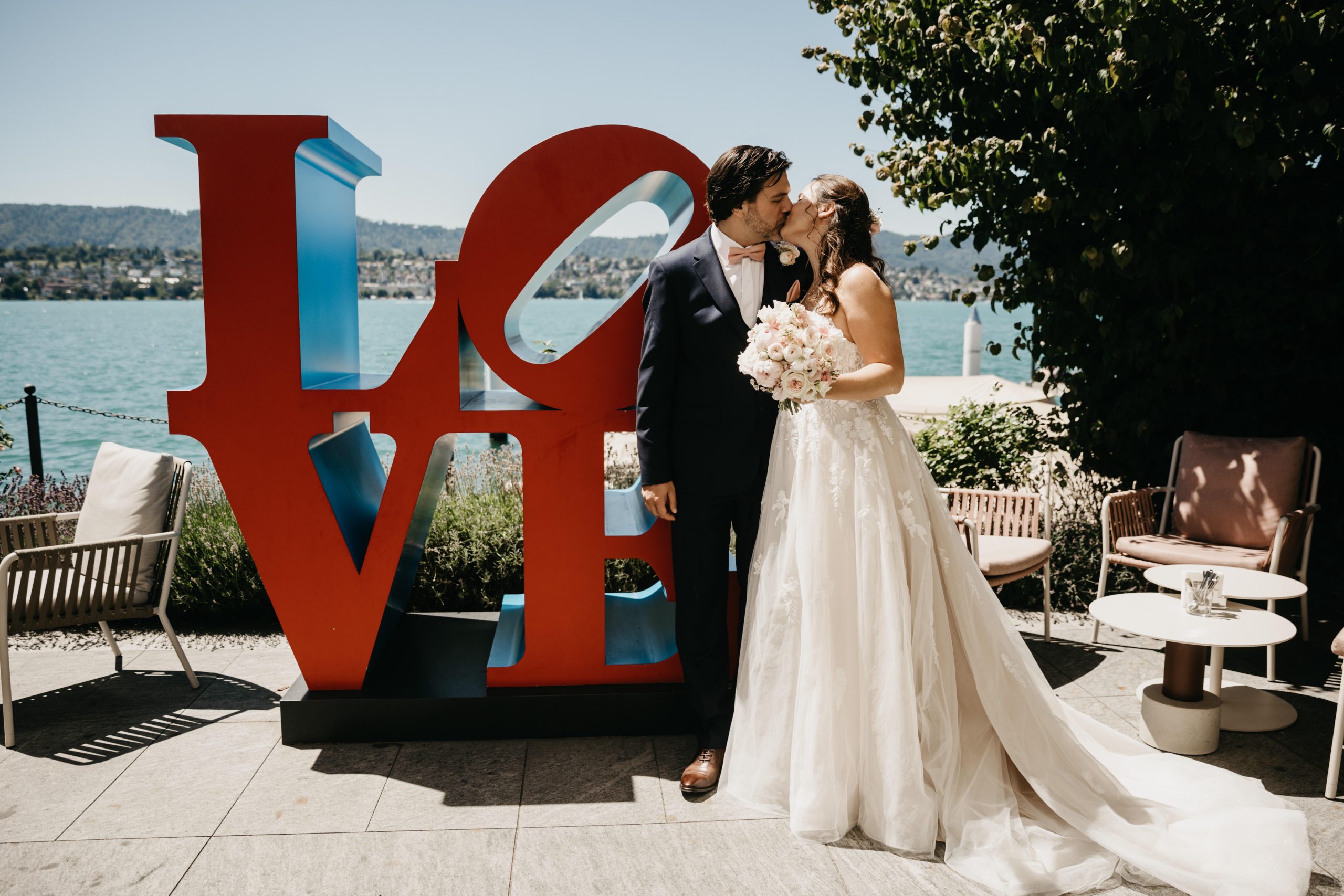 Eine Braut und ein Bräutigam stehen neben einer großen roten und blauen LOVE-Skulptur an einem See und küssen sich. Die Braut hält einen Blumenstrauß in der Hand und trägt ein weißes Kleid; der Bräutigam trägt einen dunklen Anzug. Sie sind von Sitzgelegenheiten im Freien und Bäumen umgeben.