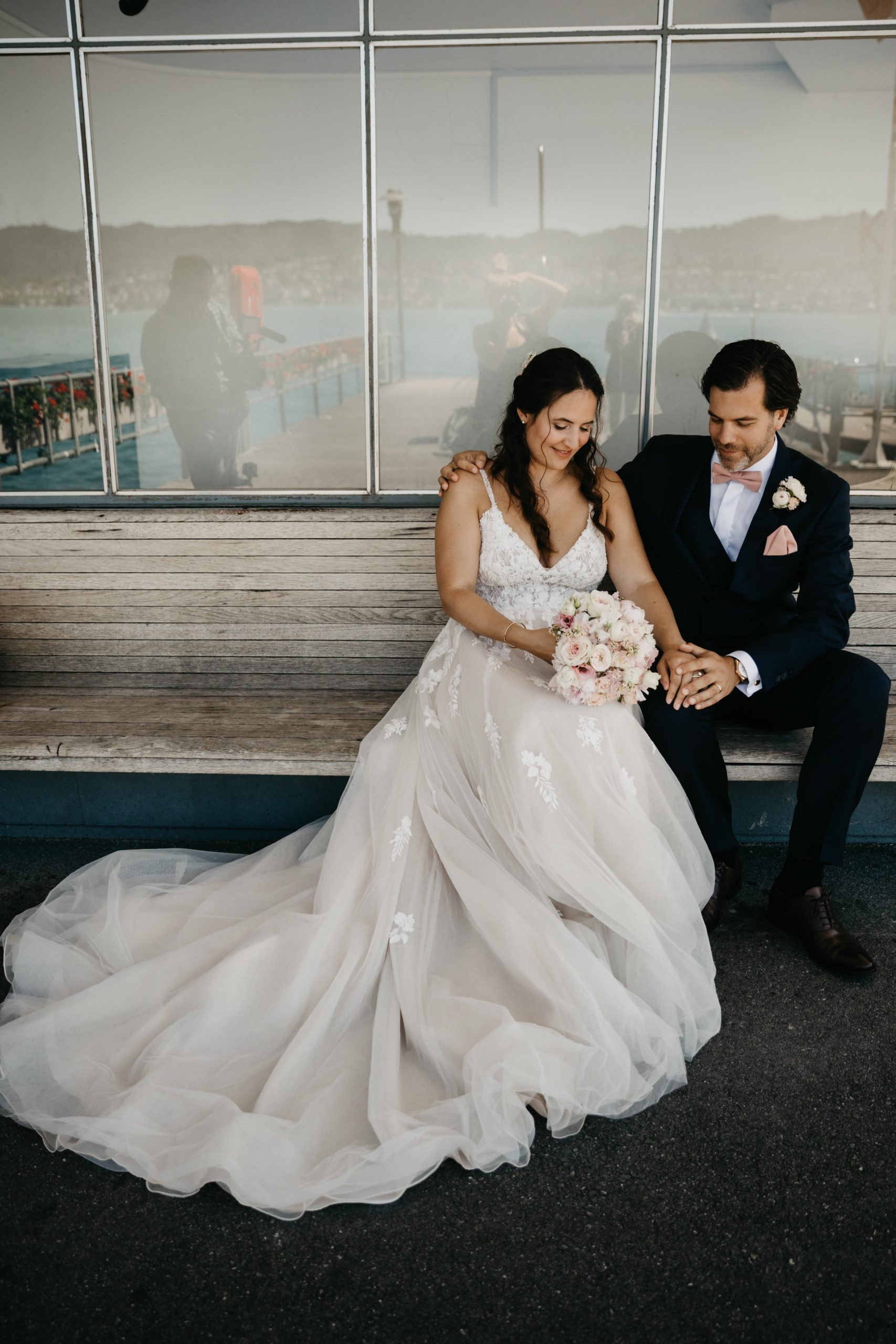 Eine Braut in einem weißen Hochzeitskleid und ein Bräutigam in einem marineblauen Anzug sitzen eng beieinander auf einer Holzbank, lächeln und halten sich an den Händen, während sich hinter ihnen ein Blumenstrauß und eine malerische Außenkulisse in Glas spiegeln.