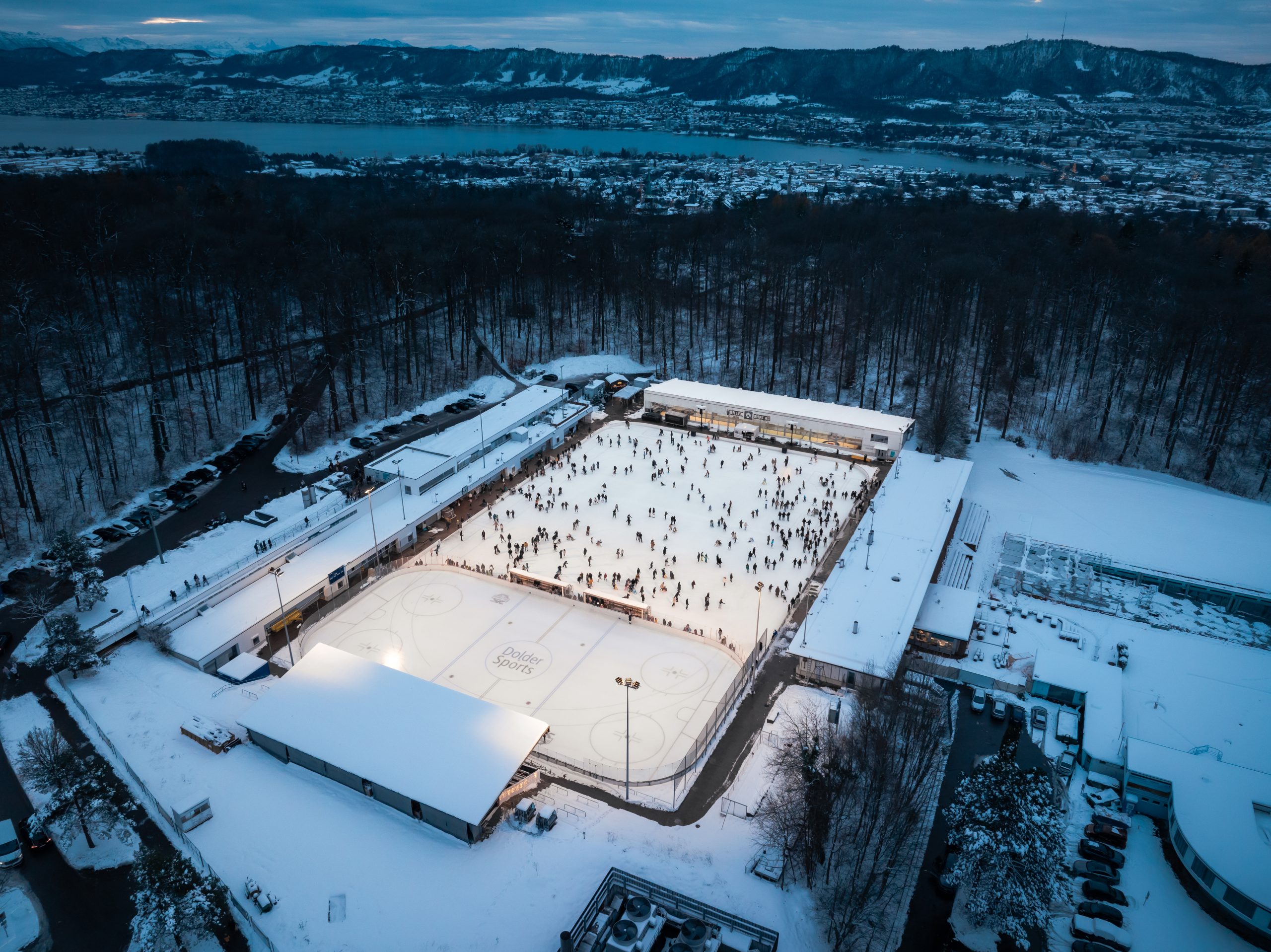 Luftaufnahme einer schneebedeckten Eislaufbahn im Freien, die ein lebhaftes Freizeitangebot bietet, da viele Menschen Schlittschuh laufen. Bäume und Gebäude begrenzen die Eisbahn, in der Ferne sind ein See und Hügel unter einem bewölkten Himmel zu sehen.