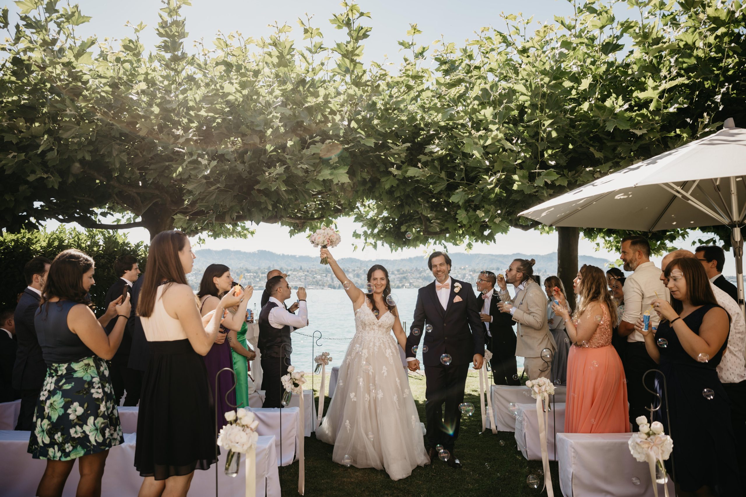 Ein frisch verheiratetes Paar schreitet lächelnd und händchenhaltend den Gang entlang, während die Gäste auf beiden Seiten stehen, klatschen und Fotos machen. Die Braut hebt feierlich ihren Brautstrauß unter dem Blätterdach der Bäume in die Höhe.