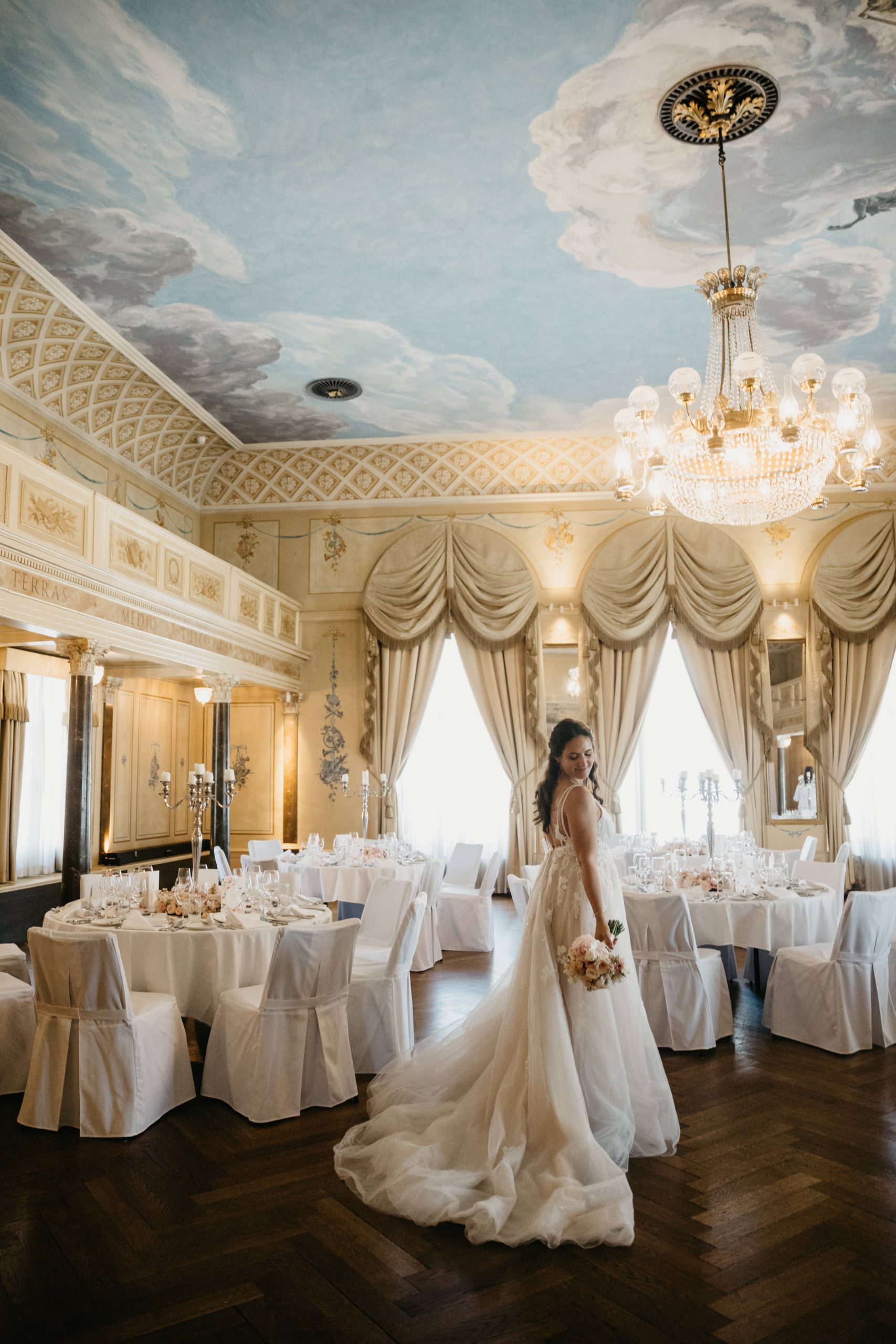 Eine Braut in einem weißen Hochzeitskleid hält einen Blumenstrauß und steht in einem eleganten, verzierten Ballsaal mit runden Tischen, drapierten Vorhängen, einer bemalten Decke und einem großen Kronleuchter.