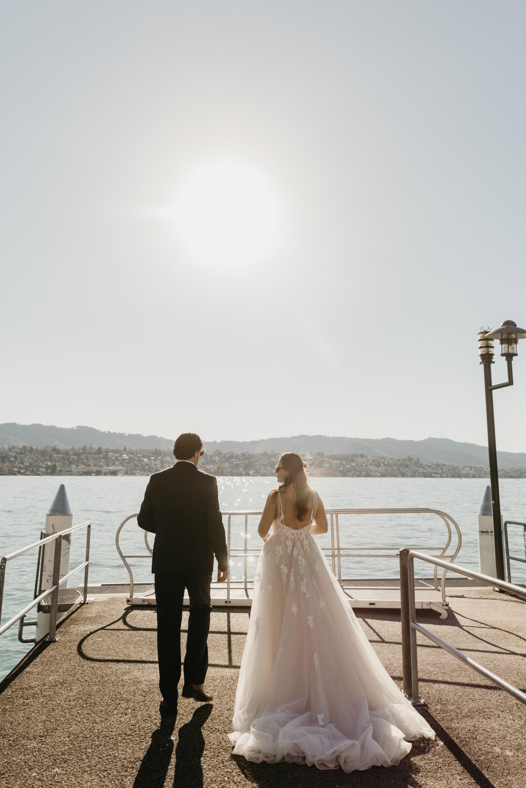 Eine Braut in einem weißen Kleid und ein Bräutigam in einem dunklen Anzug gehen gemeinsam auf einem sonnenbeschienenen Pier am Wasser spazieren, den Blick von der Kamera abgewandt in Richtung eines hellen Himmels und entfernter Hügel.