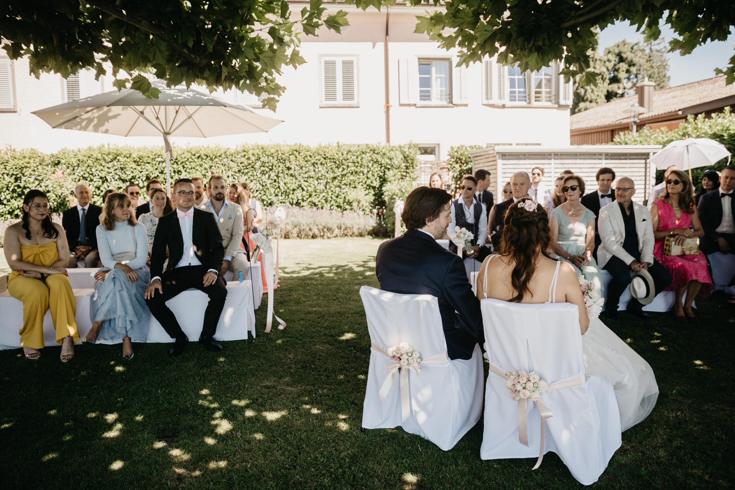 Eine Braut und ein Bräutigam sitzen ihren Hochzeitsgästen im Freien unter einem Baum gegenüber. Die formell gekleideten Gäste sitzen auf weißen Stühlen und Bänken, einige unter Sonnenschirmen, während das Sonnenlicht durch die Blätter fällt.