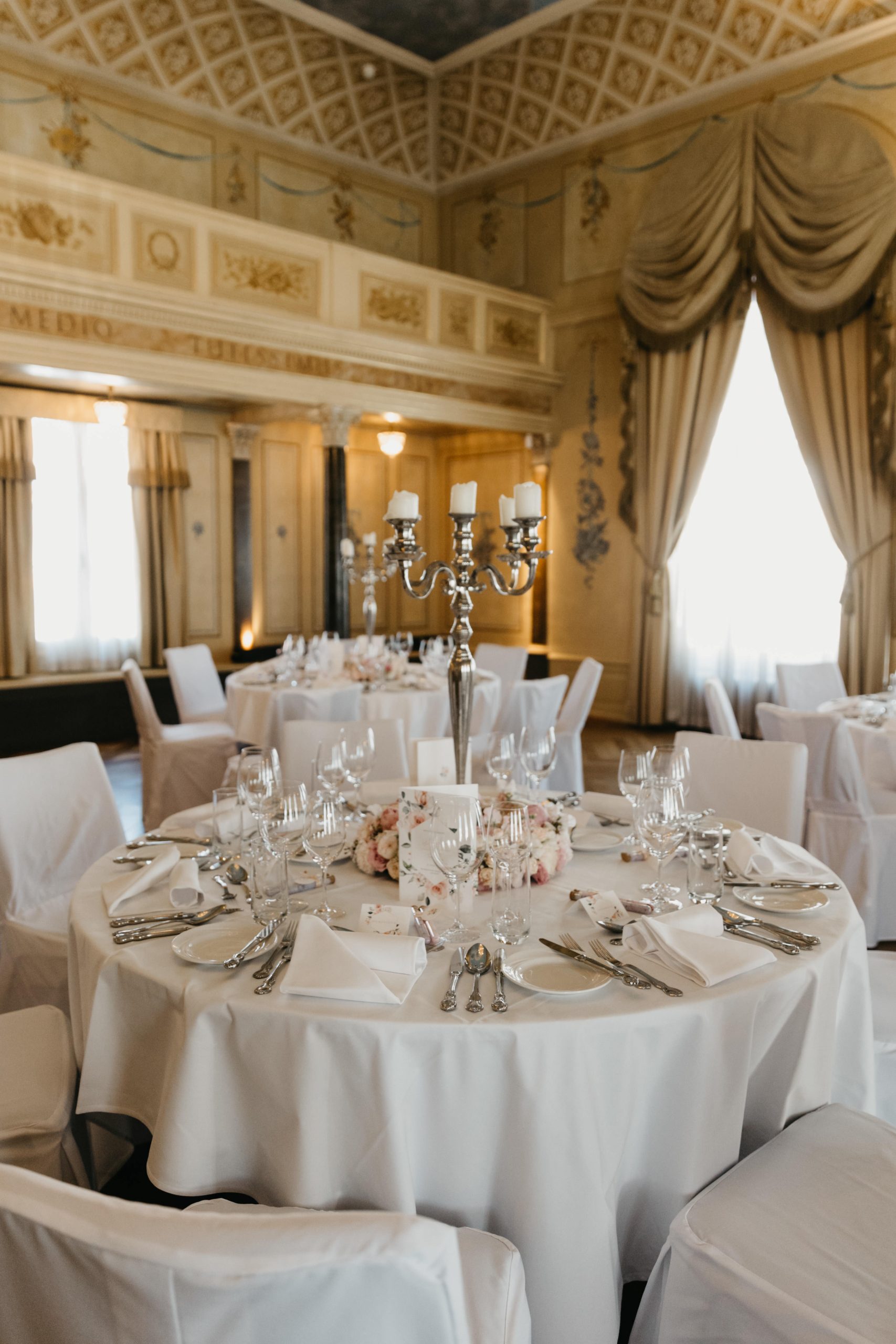 Eleganter Bankettsaal mit verzierten Wänden und Vorhängen, runden Tischen mit weißer Bettwäsche, feinem Porzellan, Gläsern, Silberbesteck, Blumenschmuck und Silberleuchtern.