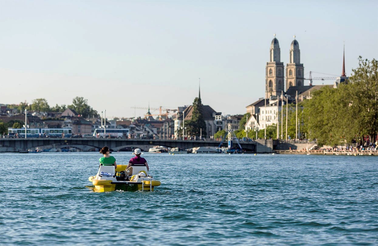 Zwei Personen genießen ein beliebtes Freizeitangebot und paddeln in einem gelben Boot auf einem ruhigen Fluss in Richtung einer Stadt mit historischen Gebäuden und einer zweitürmigen Kirche. Bäume und eine Brücke säumen die Uferpromenade unter einem klaren Himmel.