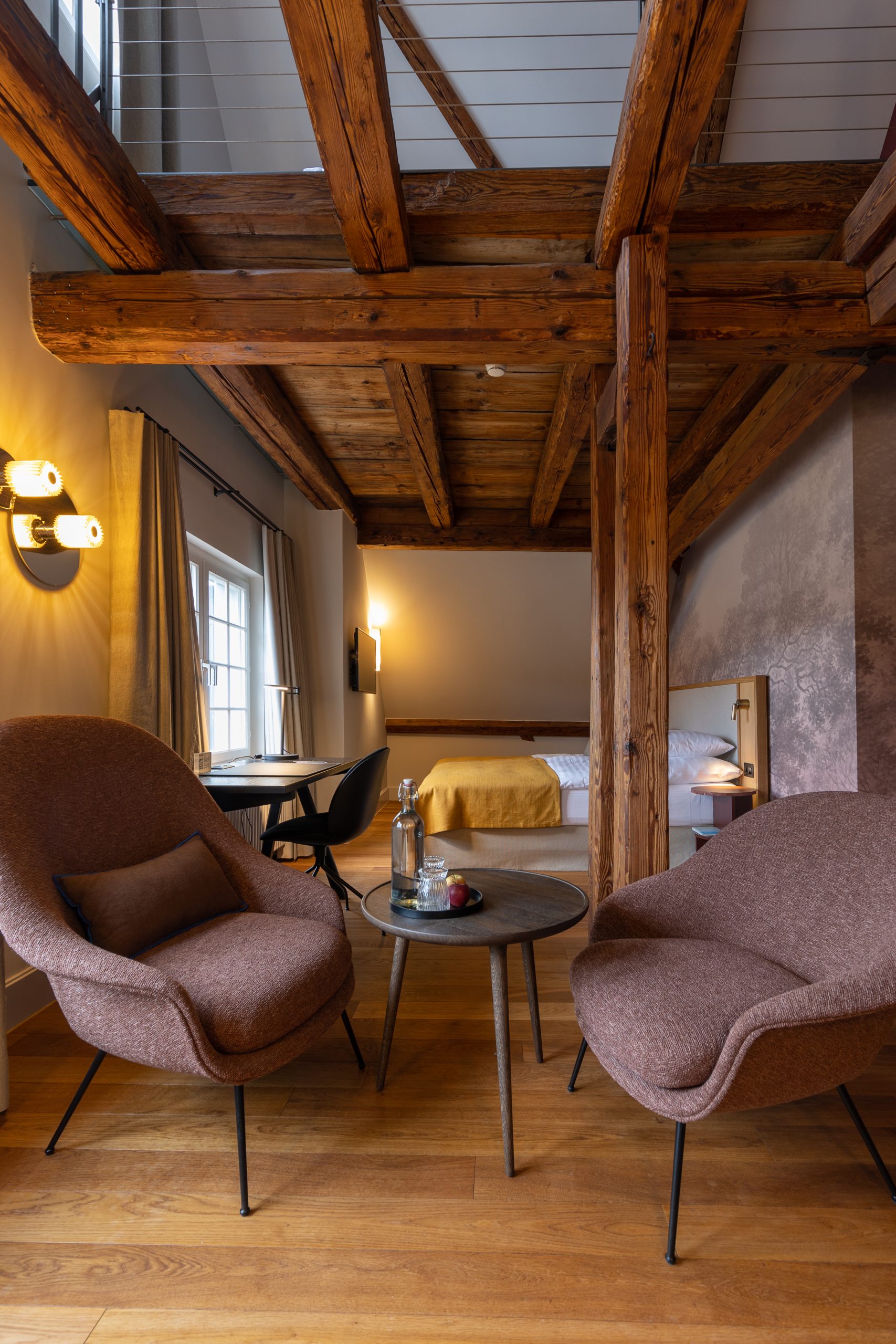 Modernes gemütliches Loft mit freiliegenden Holzbalken, zwei braunen Sesseln um einen kleinen Tisch mit Wasser und Äpfeln und einem Bett mit einer gelben Decke im Hintergrund. Genießen Sie die warme Umgebungsbeleuchtung in diesem Familienzimmer mit Seesicht.