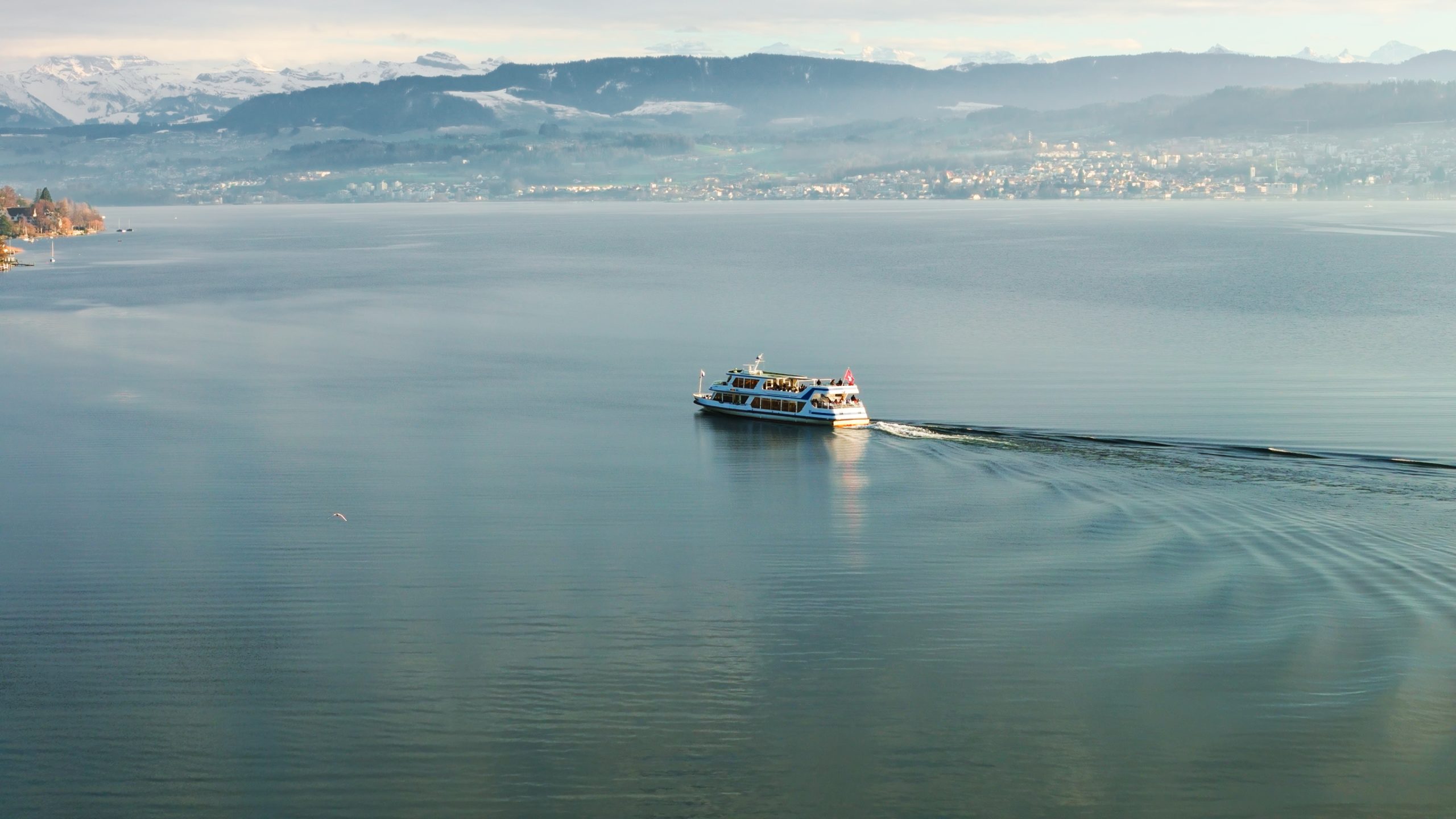 Ein weißes Passagierboot fährt über einen ruhigen See in der Nähe des Boutiquehotels Sonne und erzeugt sanftes Wellenrauschen. Unter einem wolkenverhangenen Himmel sind schneebedeckte Berge und eine Stadt zu sehen - eine ruhige Szene für diejenigen, die in der Gegend Gastronomie oder Hotel offene Stellen suchen.