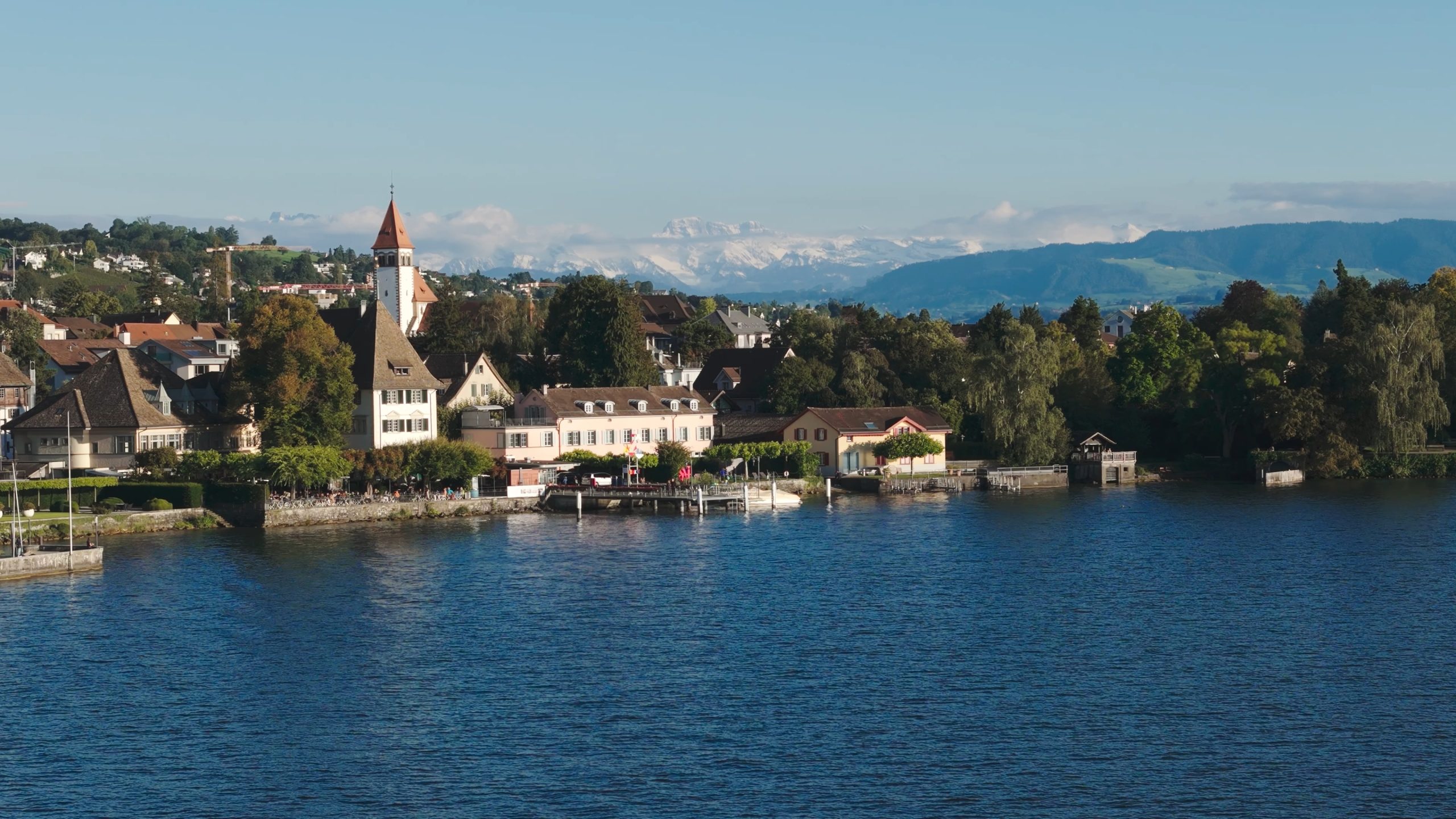Ein Dorf am See mit historischen Gebäuden, darunter eine Kirche mit hohem Turm, liegt inmitten von Bäumen und Gärten. Das nahe gelegene Boutiquehotel Sonne bietet einen atemberaubenden Blick auf den ruhigen blauen See und die Berge, die sich unter einem klaren Himmel erheben.