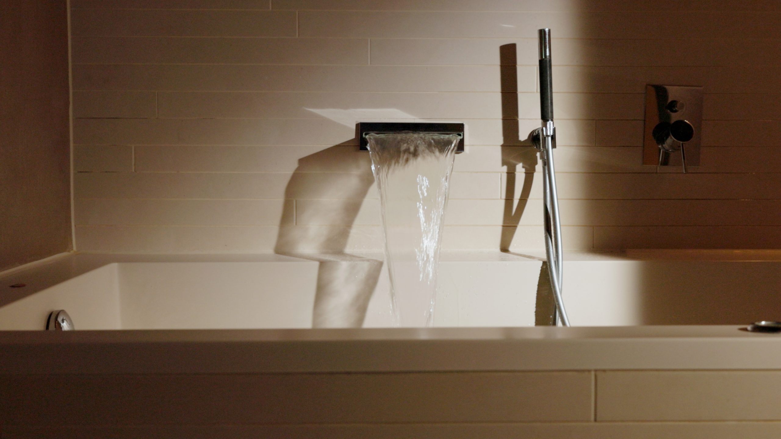 Eine moderne Badewanne mit einem breiten, flachen Wasserhahn, aus dem das Wasser in die Wanne fließt, daneben eine elegante Handbrause und Bedienelemente an einer gefliesten Wand - perfekt zum Entspannen nach einem Tag in der Hotellerie im Boutiquehotel Sonne.