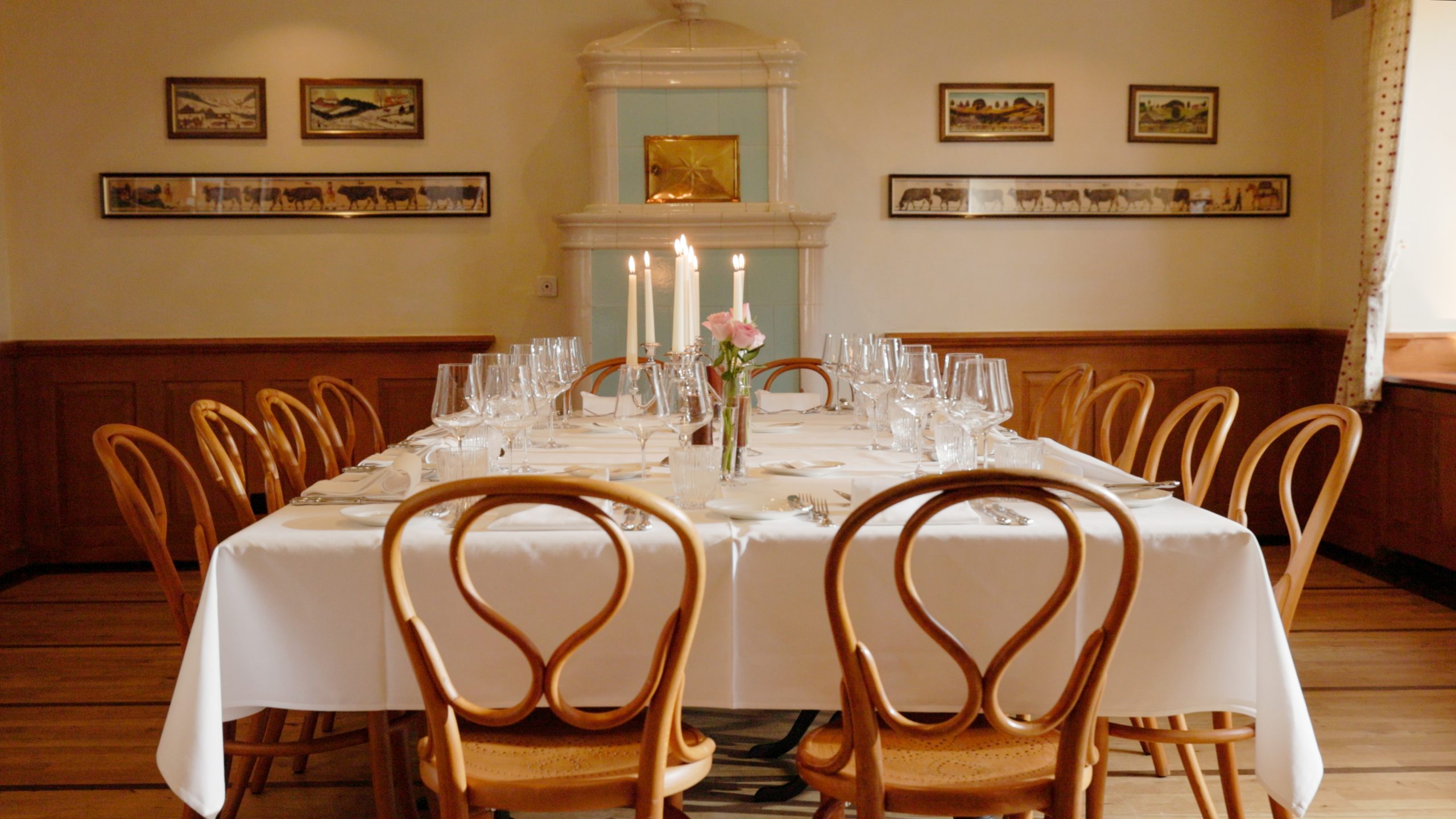 Ein Esstisch für acht Personen mit weißer Tischdecke, Weingläsern, Tellern und Besteck erwartet Sie im warm beleuchteten Boutiquehotel Sonne. Drei brennende Kerzen und rosafarbene Blumen vervollständigen die gemütliche Atmosphäre - ideal für alle, die einen Hotellerie-Job oder einen Gastrojob suchen.