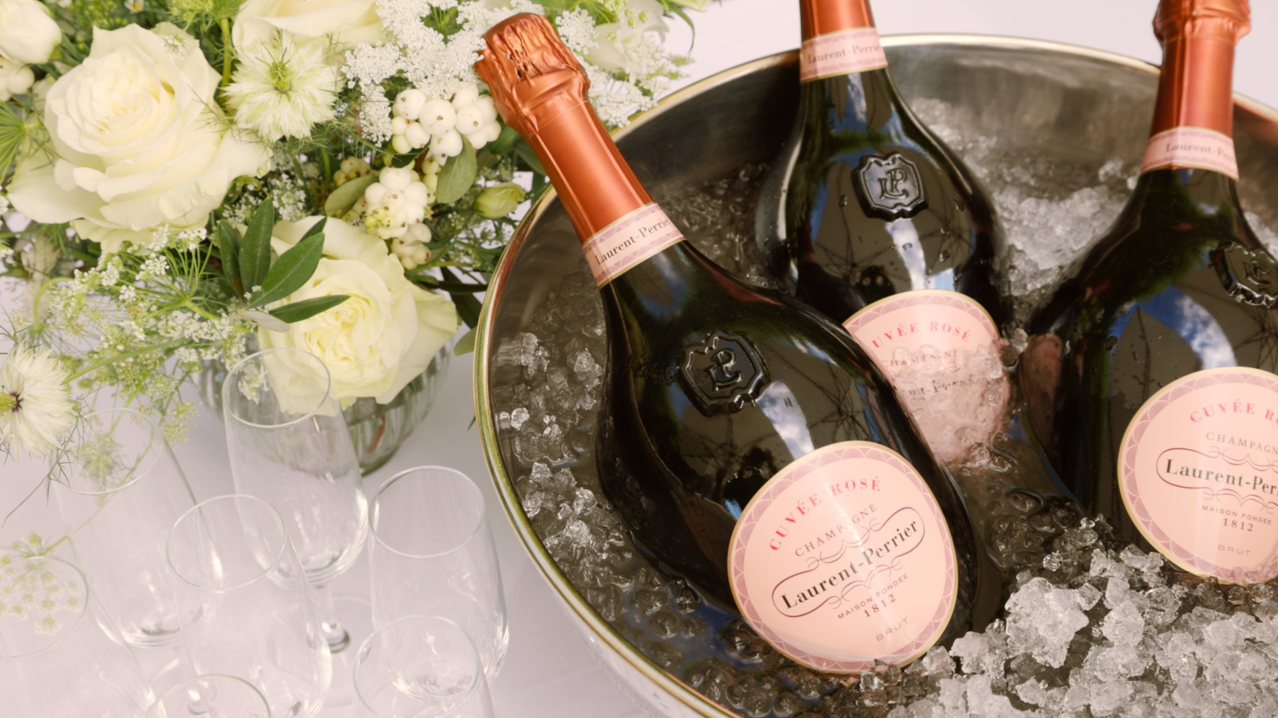 Zwei Flaschen Laurent-Perrier Cuvée Rosé Champagner, die in einem mit Eis gefüllten Eimer kühlen, zusammen mit einem Strauß weißer Blumen und mehreren leeren Gläsern - perfekt, um neue Hotellerie-Jobs oder Hotel offene Stellen zu feiern.