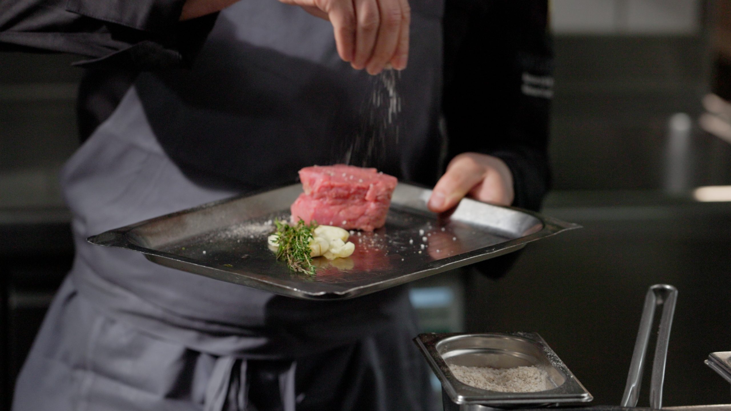 Ein Koch im Hotel Sonne streut Salz auf ein rohes Steak auf einem Metalltablett, begleitet von Knoblauchzehen und frischen Kräutern, in einer Küchenzeile.