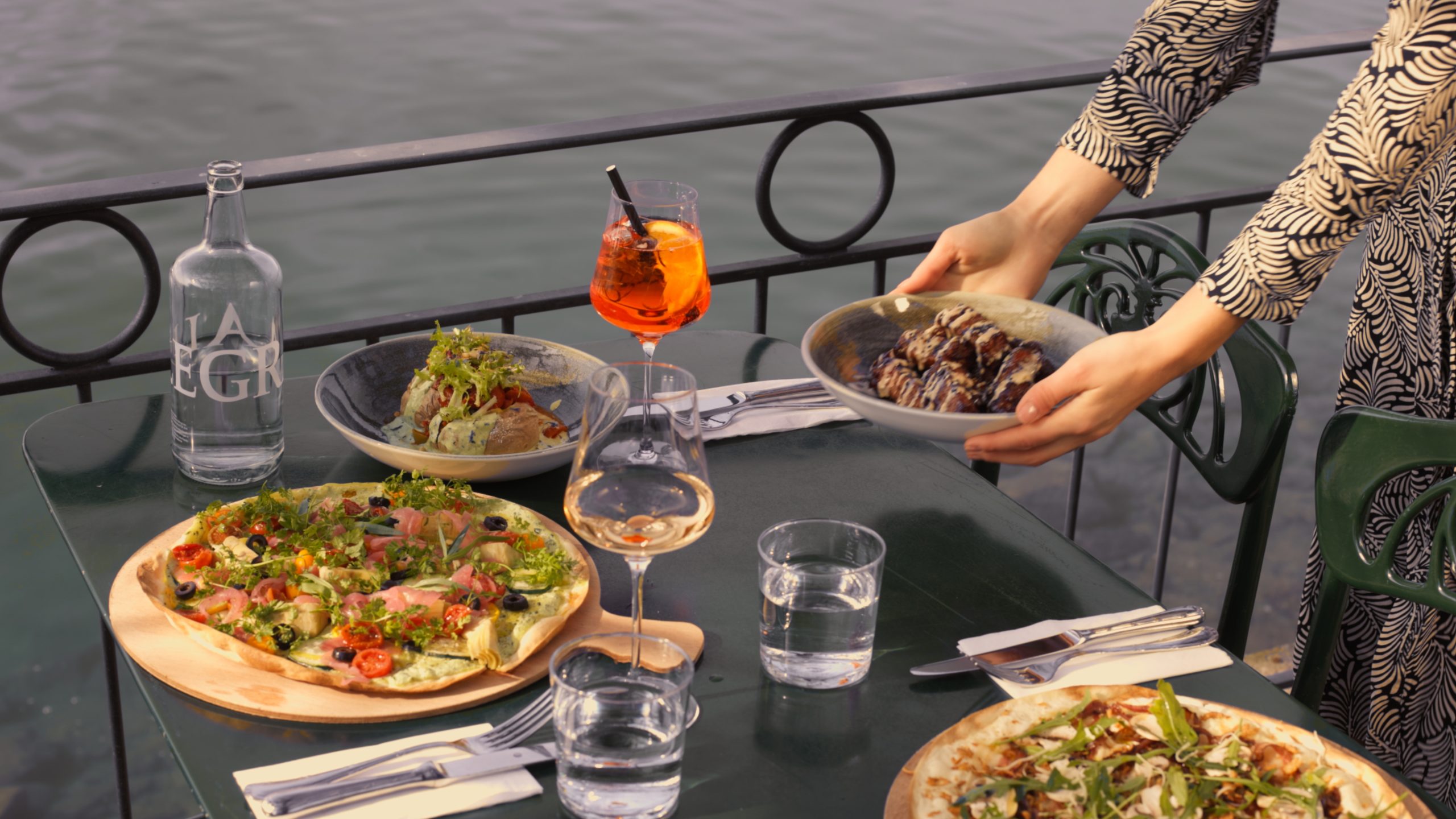 Eine Person stellt einen Teller mit gegrillten Auberginen auf einen Tisch im Freien am Wasser, der bereits mit Salaten, Pizzen, Getränken und Gläsern gefüllt ist. So entsteht eine malerische Szene im Freien im Boutiquehotel Sonne - die perfekte Inspiration für Gastrojobs.