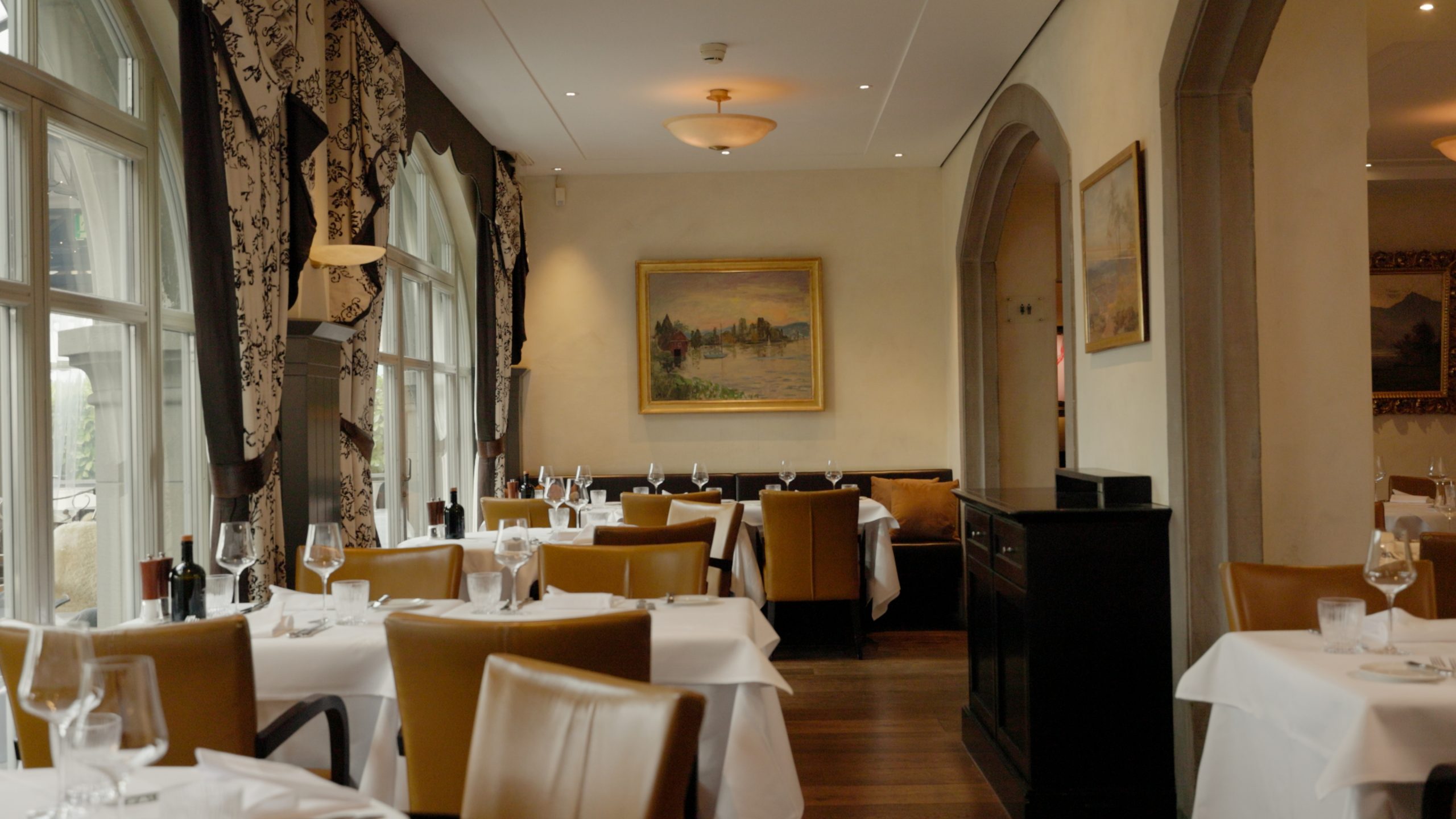 Elegantes Restaurantinterieur mit weißen Tischdecken, hellbraunen Lederstühlen und großen Fenstern. Gerahmte Landschaftsgemälde tragen zur einladenden Atmosphäre bei - perfekt für alle, die einen Hotellerie-Job suchen oder Gastrojobs ausprobieren möchten.