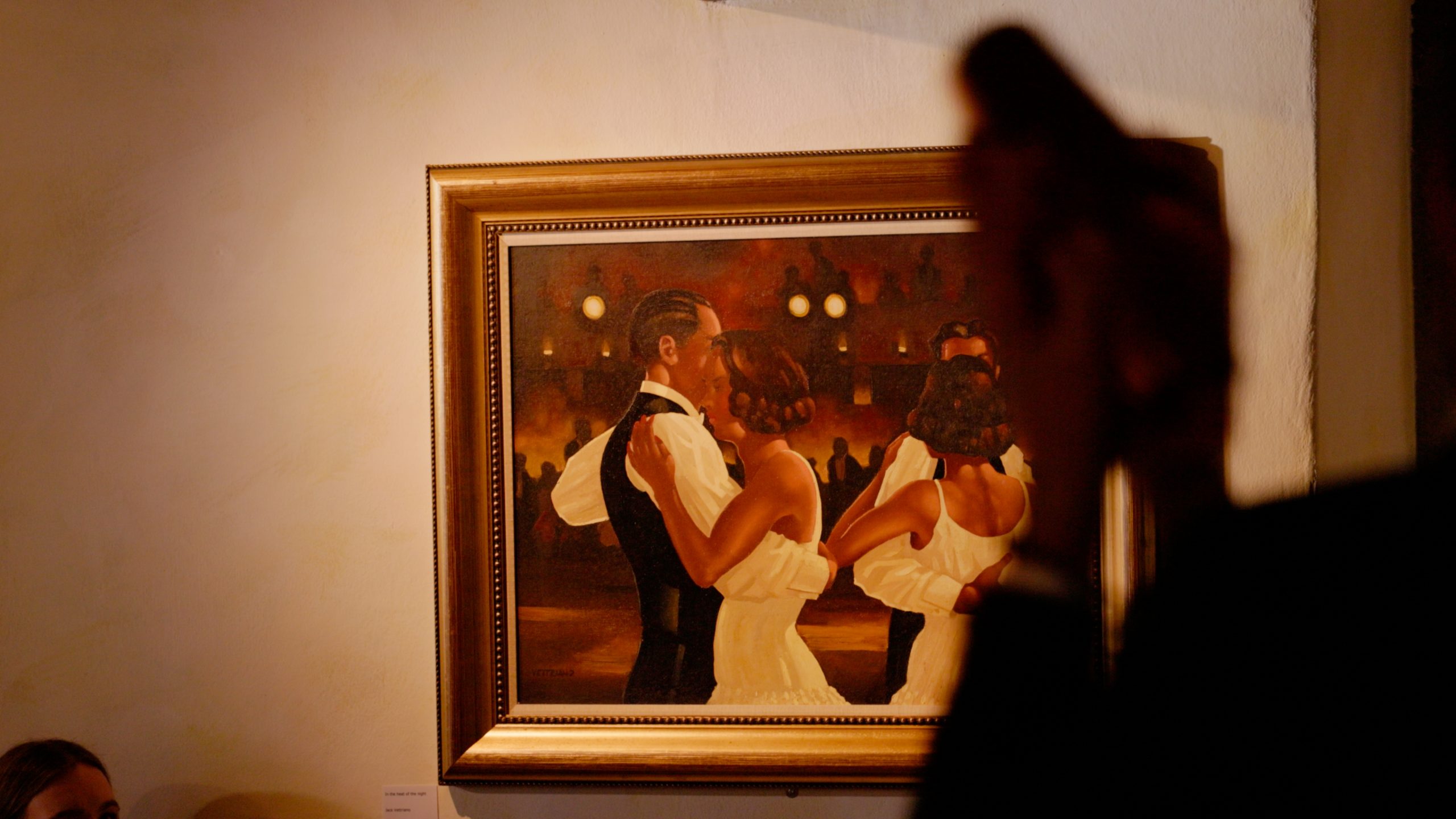 Ein gerahmtes Gemälde mit tanzenden Paaren hängt an einer Wand, die warm beleuchtet ist. Im Vordergrund ist teilweise eine schemenhafte Figur im Profil zu sehen, die einen Kontrast zwischen realen Menschen und Kunst schafft - ähnlich wie die lebhaften Szenen in der Hotellerie Jobs.