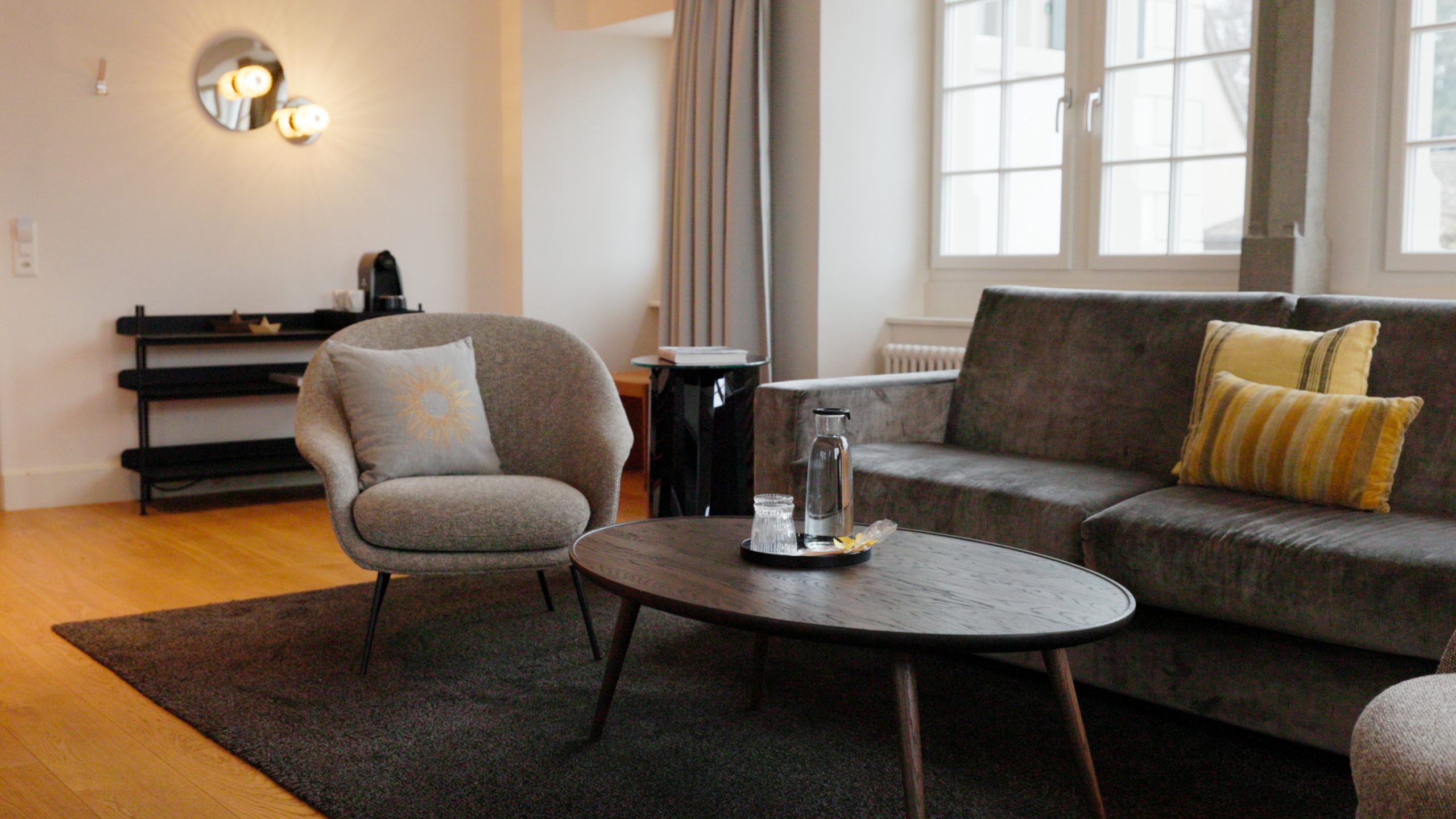 Das moderne Wohnzimmer im Boutiquehotel Sonne ist mit einem grauen Sofa, einem Sessel mit Blumenmuster, einem runden Couchtisch mit Karaffe, Holzböden, großen Fenstern sowie einem schwarzen Regal und einer Wandleuchte ausgestattet - ein einladender Raum für Gäste oder diejenigen, die in der Hotellerie arbeiten möchten.