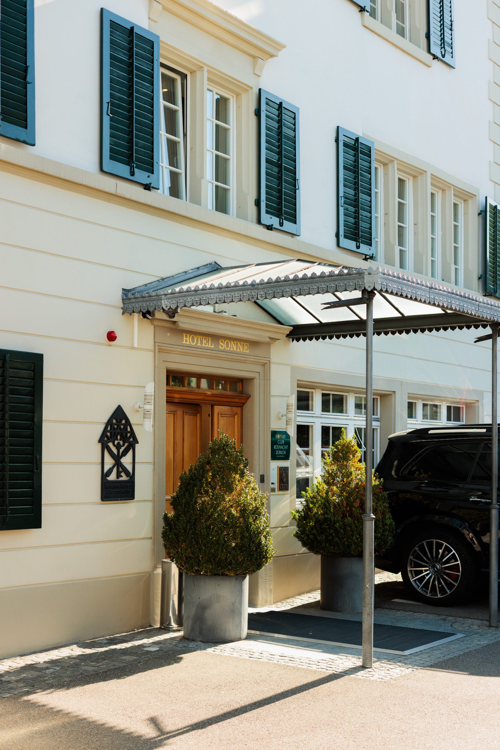 Hoteleingang mit grünen Fensterläden, einer Holztür, einer Glasmarkise und zwei großen Topfpflanzen. In der Nähe ist ein schwarzes Auto geparkt. Auf dem Schild über dem Eingang steht "Hotel Sonne".