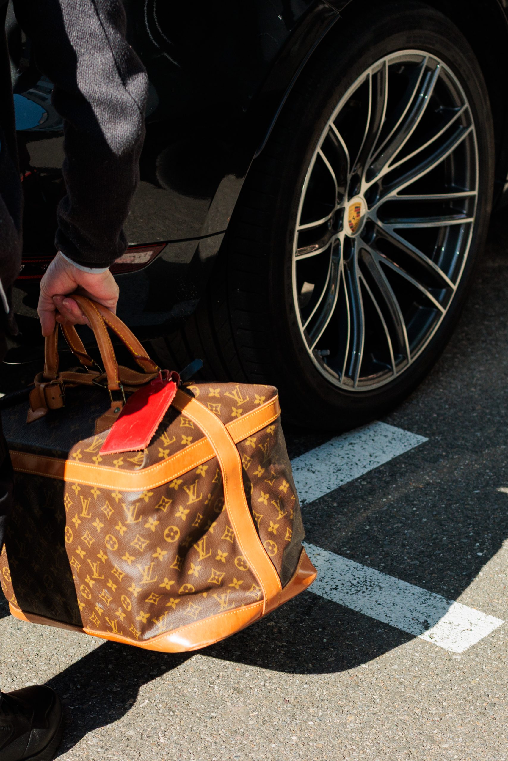 Eine Person mit einer Louis Vuitton Reisetasche neben einem schwarzen Auto mit einem sichtbaren Porsche-Rad, das auf einem sonnigen Parkplatz geparkt ist.