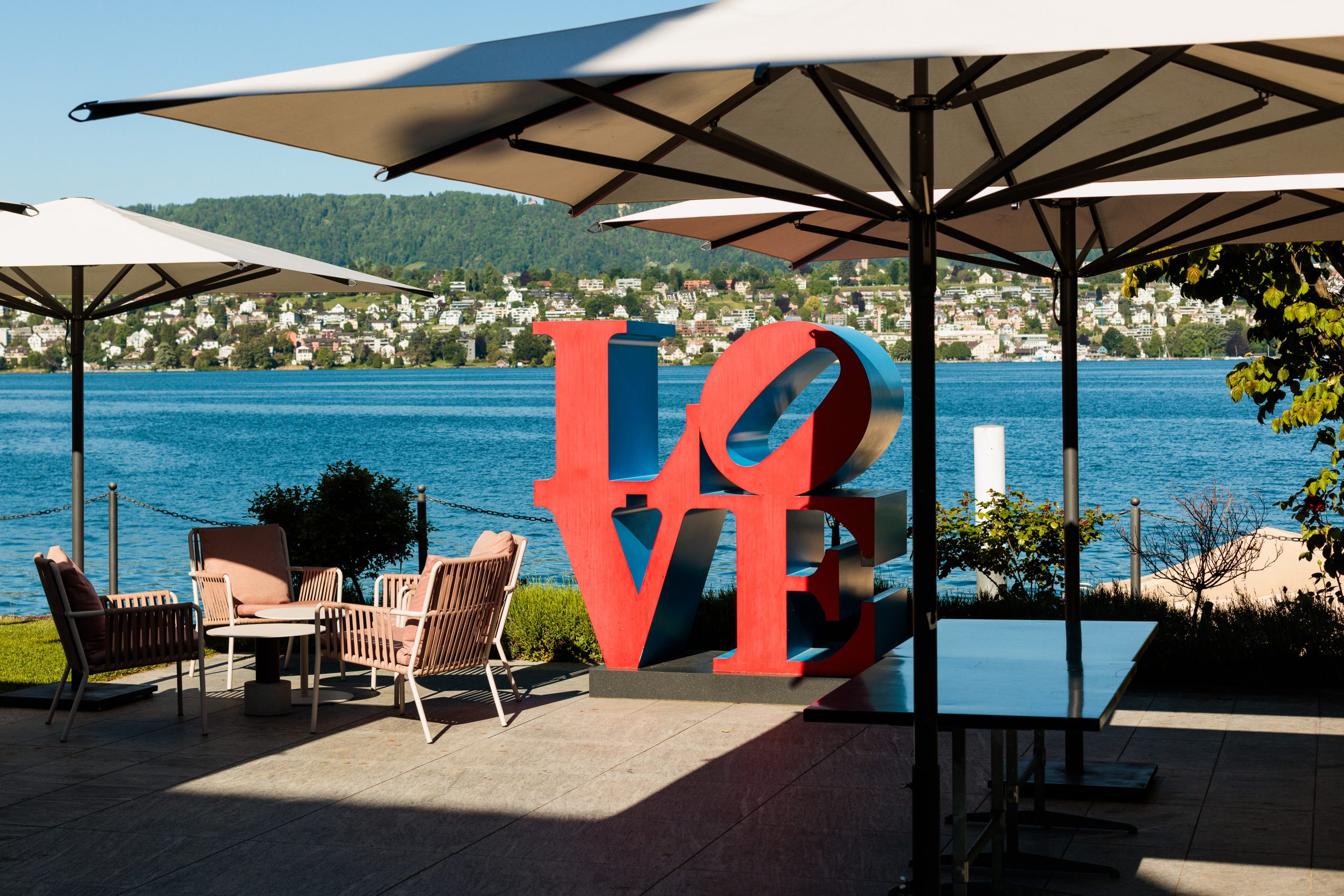 Eine große rote LOVE-Skulptur steht auf einer Terrasse in der Nähe eines Sees, umgeben von Liegestühlen und Tischen unter großen Sonnenschirmen, mit einer Stadt in den Bergen im Hintergrund.