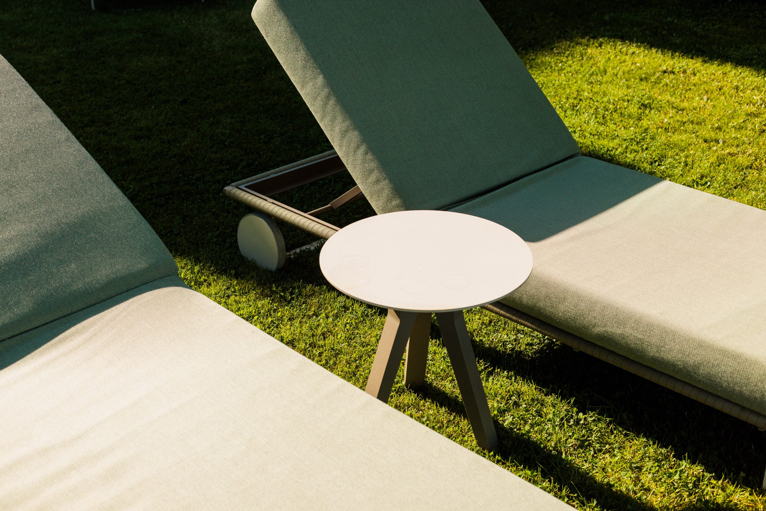 Zwei leere Liegestühle stehen auf grünem Gras, dazwischen ein kleiner runder weißer Tisch, der teilweise im Sonnenlicht liegt.
