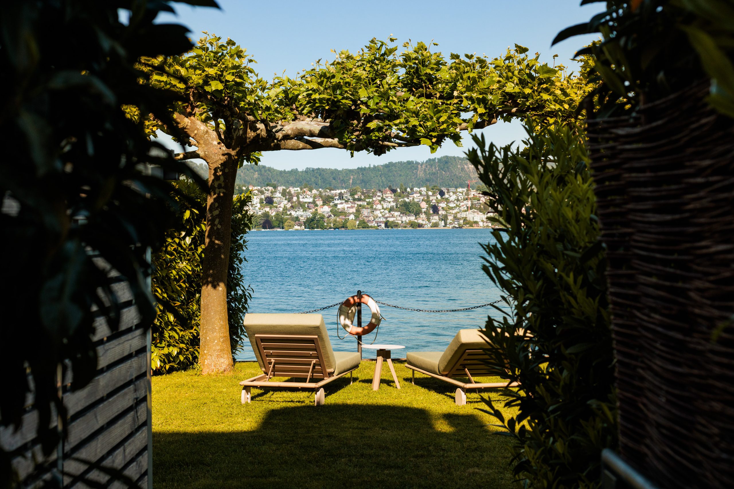 Zwei Liegestühle und ein kleiner Tisch stehen im Gras unter Bäumen an einem See, eine Person beugt sich über das Wasser, und auf der anderen Seite des Sees ist eine malerische Stadt unter einem klaren blauen Himmel zu sehen.