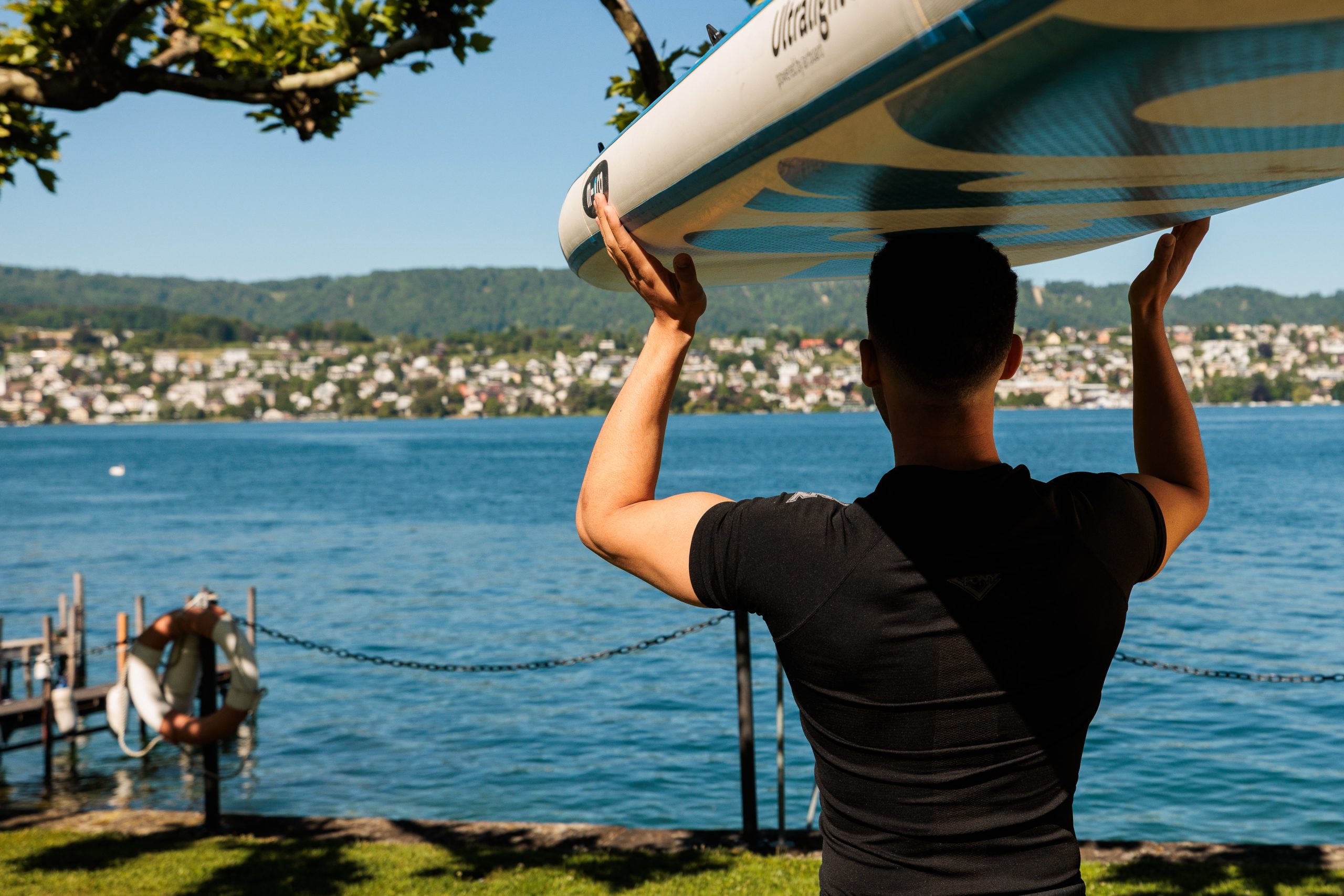 A person carries a paddleboard on their head near a calm lake, with a town and green hills visible across the water under a clear, sunny sky.