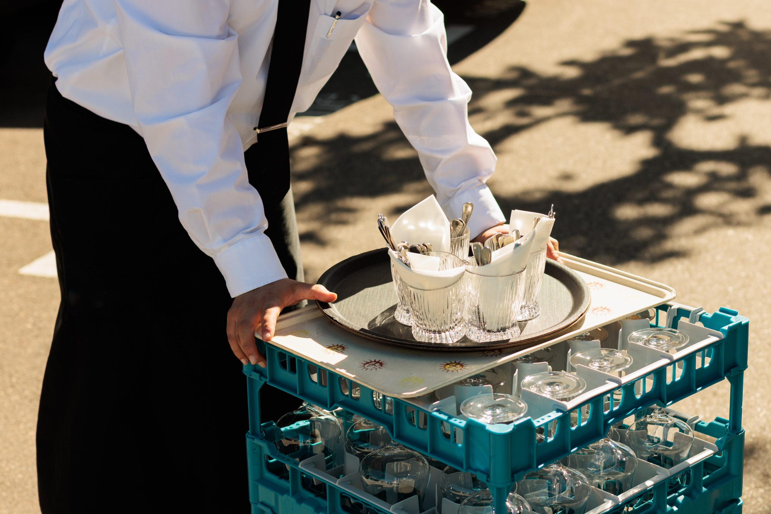Eine Person in weißem Hemd und schwarzer Krawatte trägt ein Tablett mit Gläsern und Besteck und stellt es auf ein Gestell mit sauberen Gläsern im Freien im Sonnenlicht.