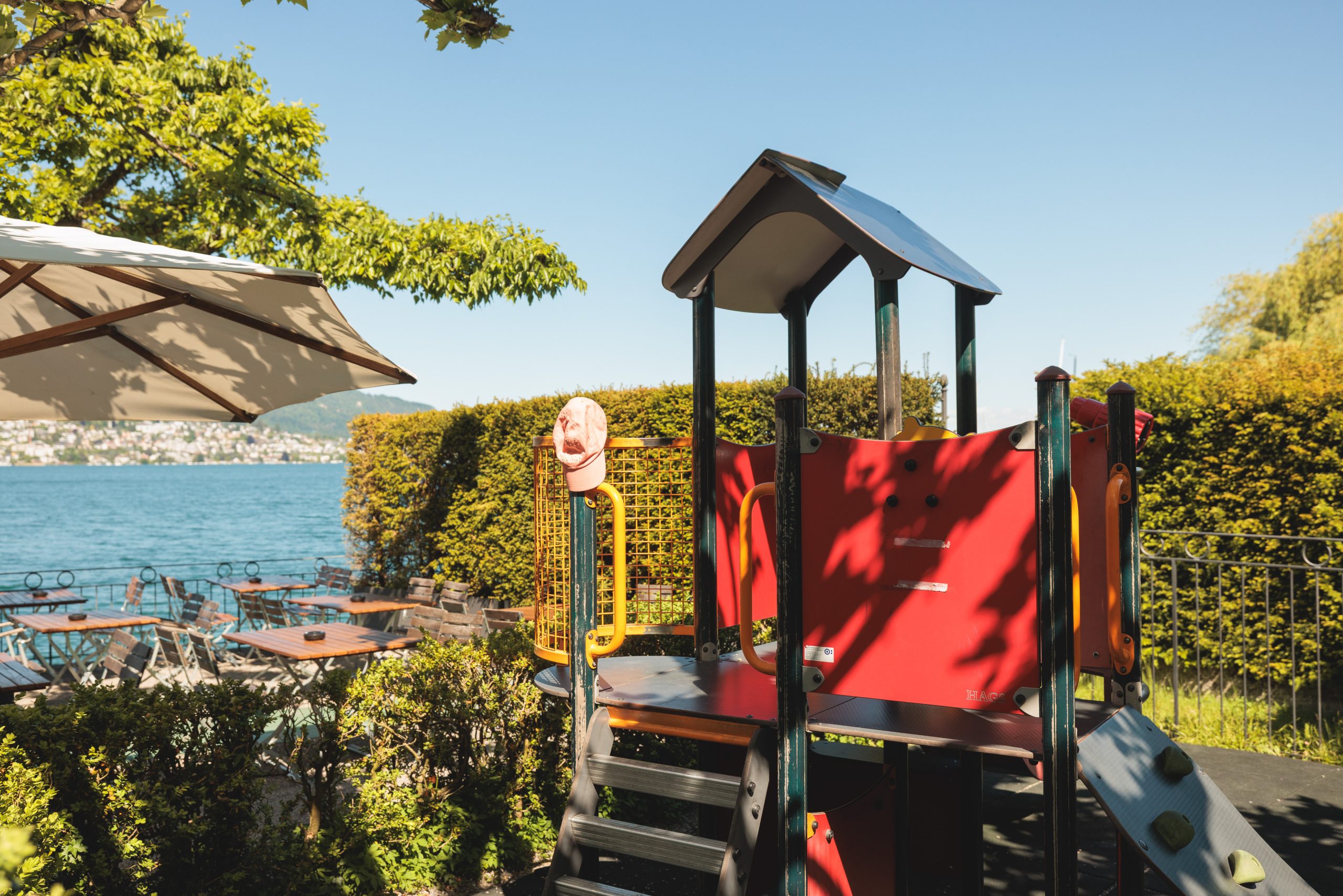Ein kleines Spielgerät mit Stufen und einer Rutsche steht neben einer Caféterrasse am See, umgeben von grünen Büschen und Bäumen unter einem klaren blauen Himmel. Tische und Stühle sind in der Nähe des Wassers im Hintergrund zu sehen.