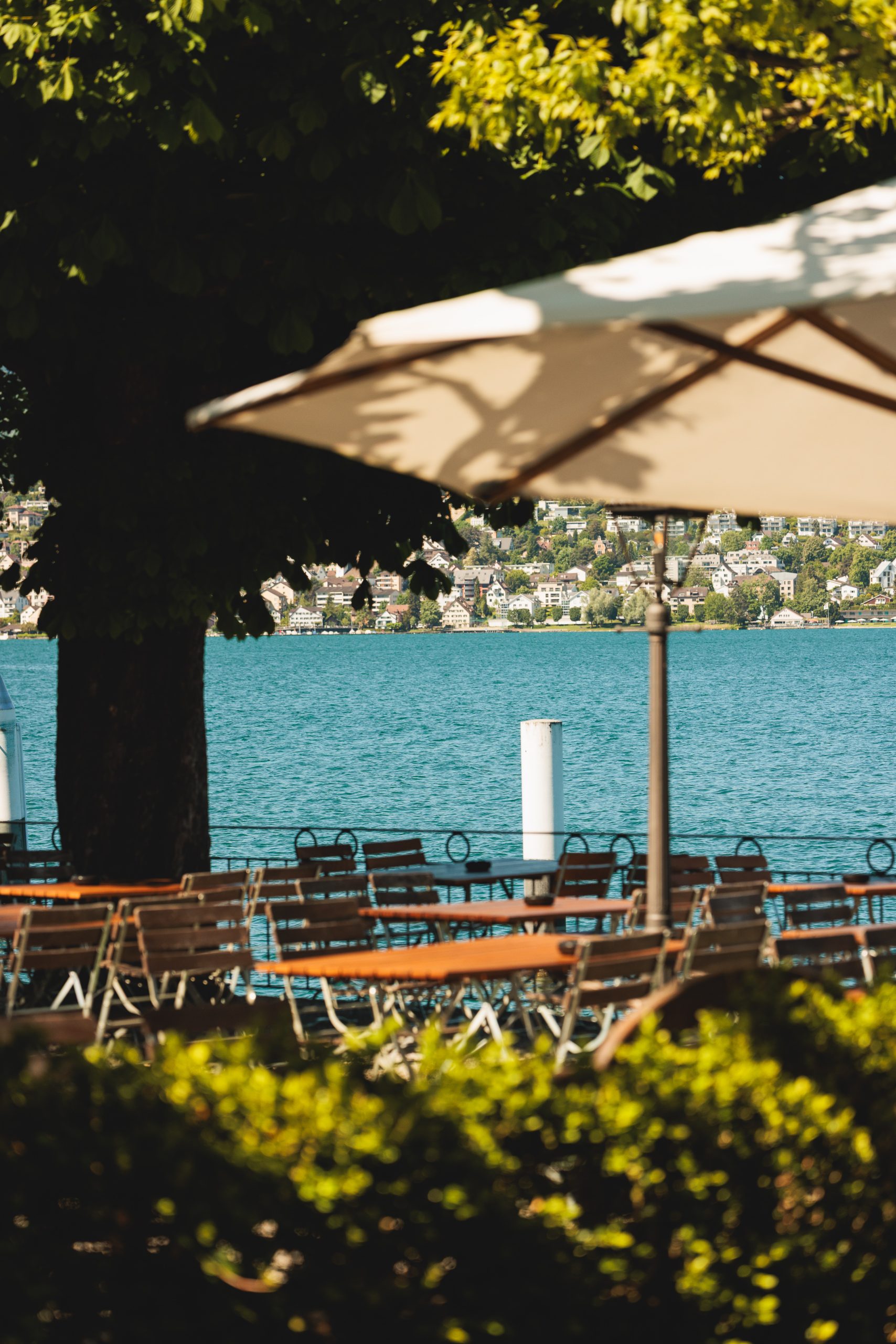 Ein Außencafé mit leeren Holztischen und Stühlen unter einem großen Sonnenschirm, mit Blick auf einen blauen See mit Häusern am gegenüberliegenden Ufer; die Umgebung ist grün und sonnig.