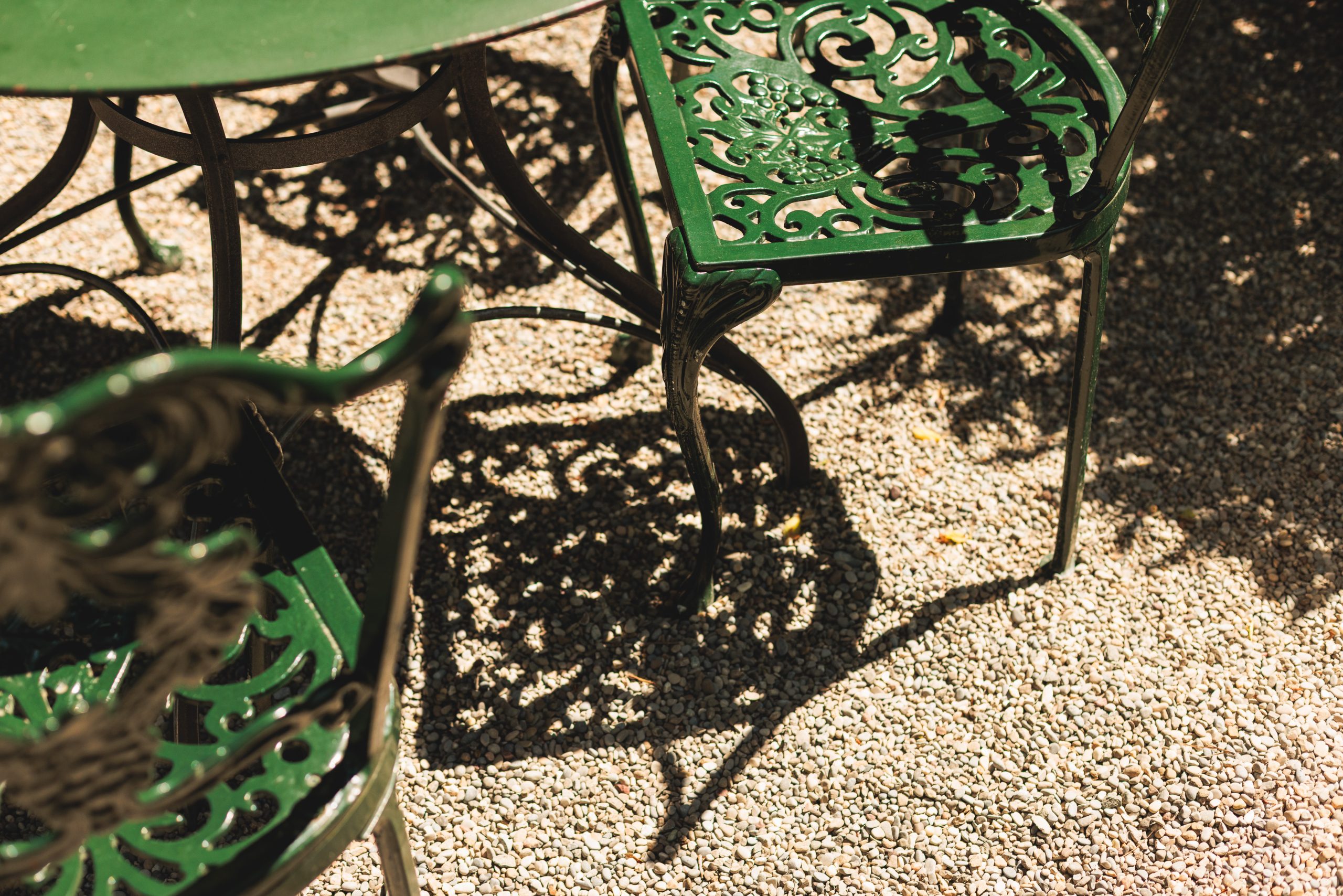 Ein grüner schmiedeeiserner Tisch und Stühle werfen im hellen Sonnenlicht komplizierte Schatten auf eine Kiesfläche. Die kunstvollen Metallarbeiten und die Textur des Kieses sind deutlich zu erkennen.