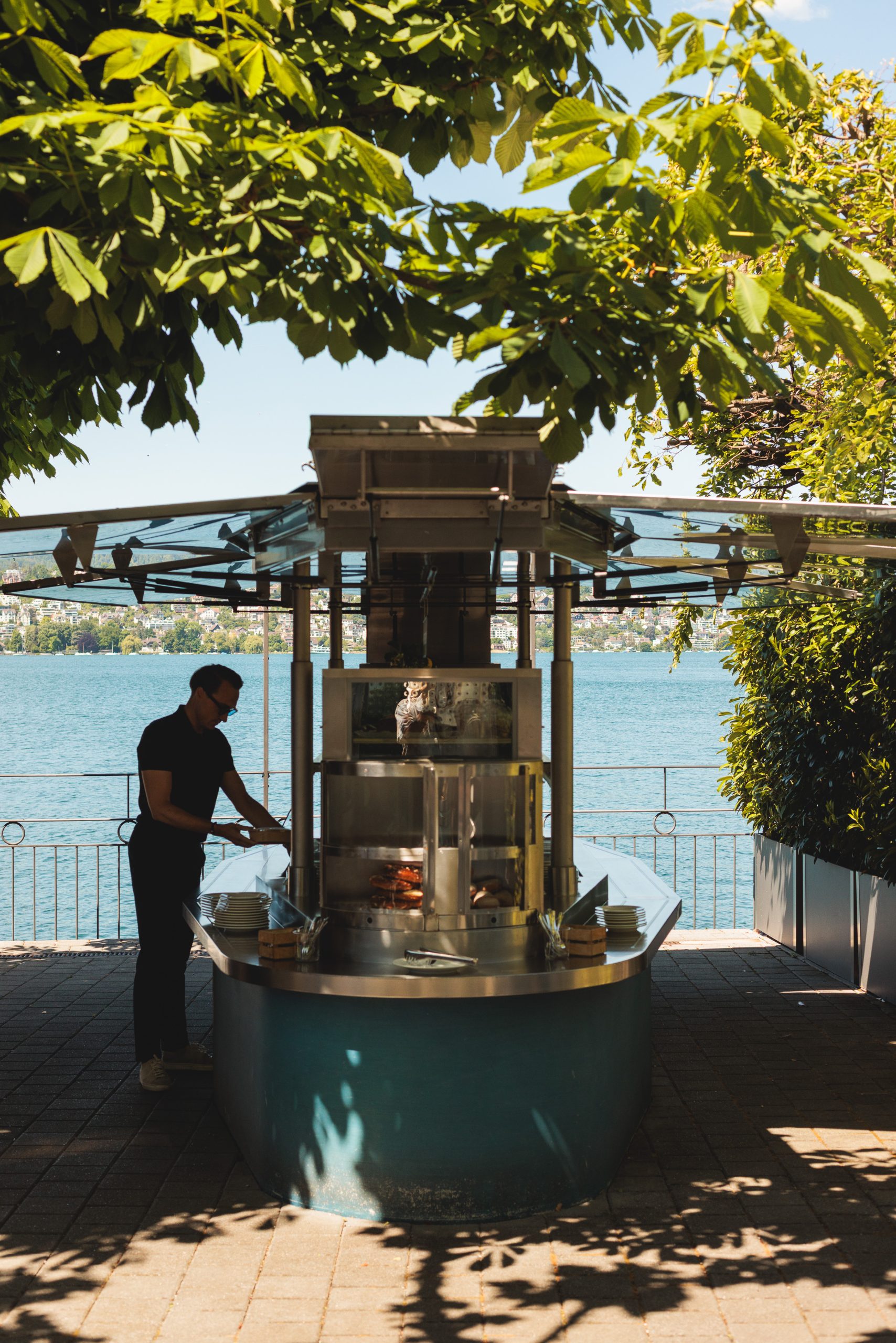 Eine Person bereitet an einem sonnigen Tag an einem Kiosk im Freien unter grünen Bäumen, mit einem See und entfernten Gebäuden im Hintergrund, ein Essen zu.