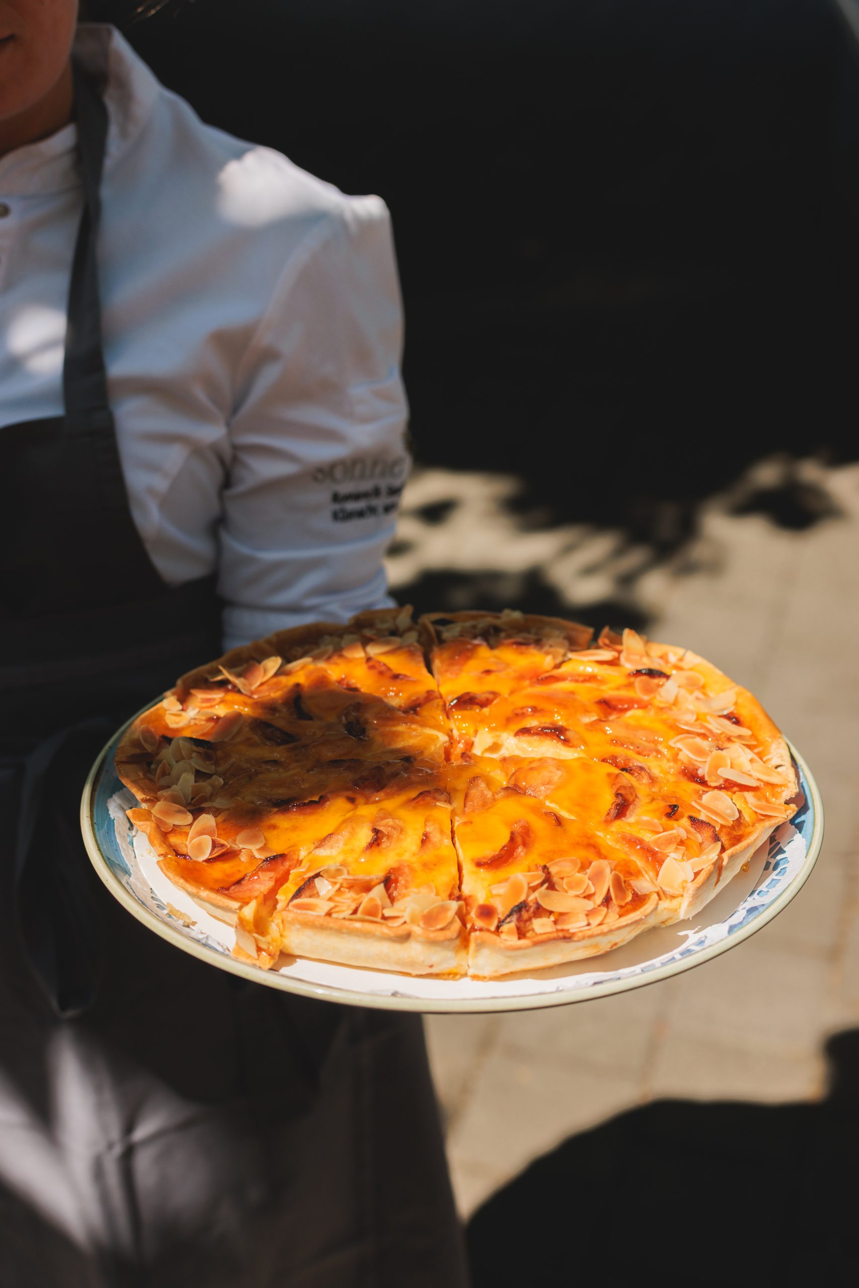Eine Person in weißer Kochjacke und dunkler Schürze hält einen Teller mit einer aufgeschnittenen Torte, die mit Mandeln und einem glänzenden orangefarbenen Belag bedeckt ist, und steht im Freien im Sonnenlicht.