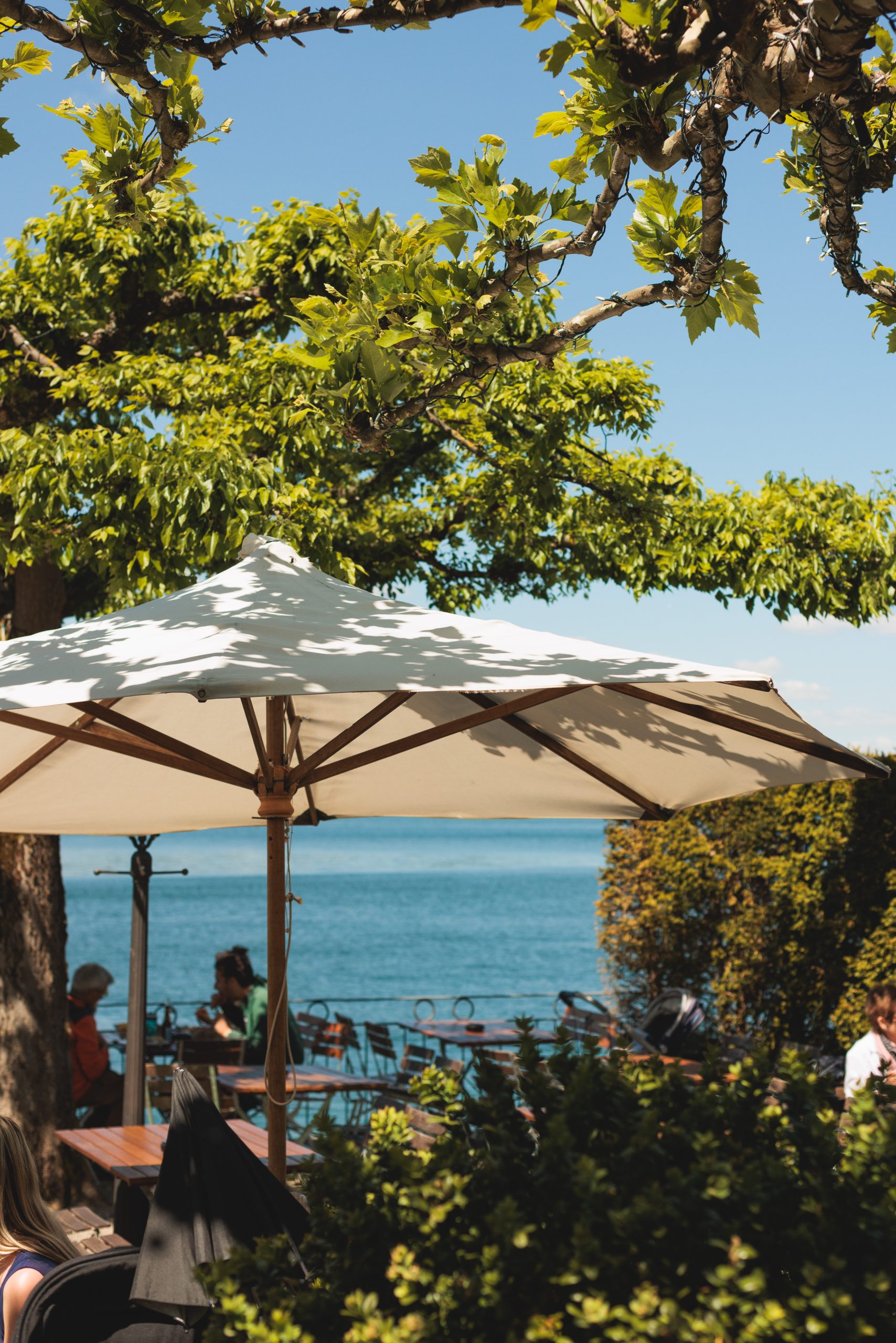Ein weißer Sonnenschirm beschattet ein Straßencafé mit Holztischen und Stühlen, umgeben von Grün und mit Blick auf einen blauen See unter klarem Himmel.