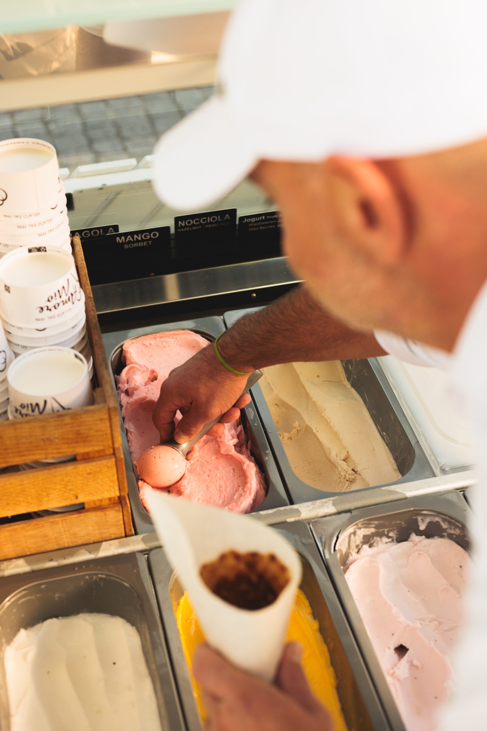 Eine Person mit weißer Mütze, die an einem Eisstand rosa Eis in eine Waffel schöpft, wobei verschiedene Geschmacksrichtungen und Pappbecher in der Vitrine zu sehen sind.