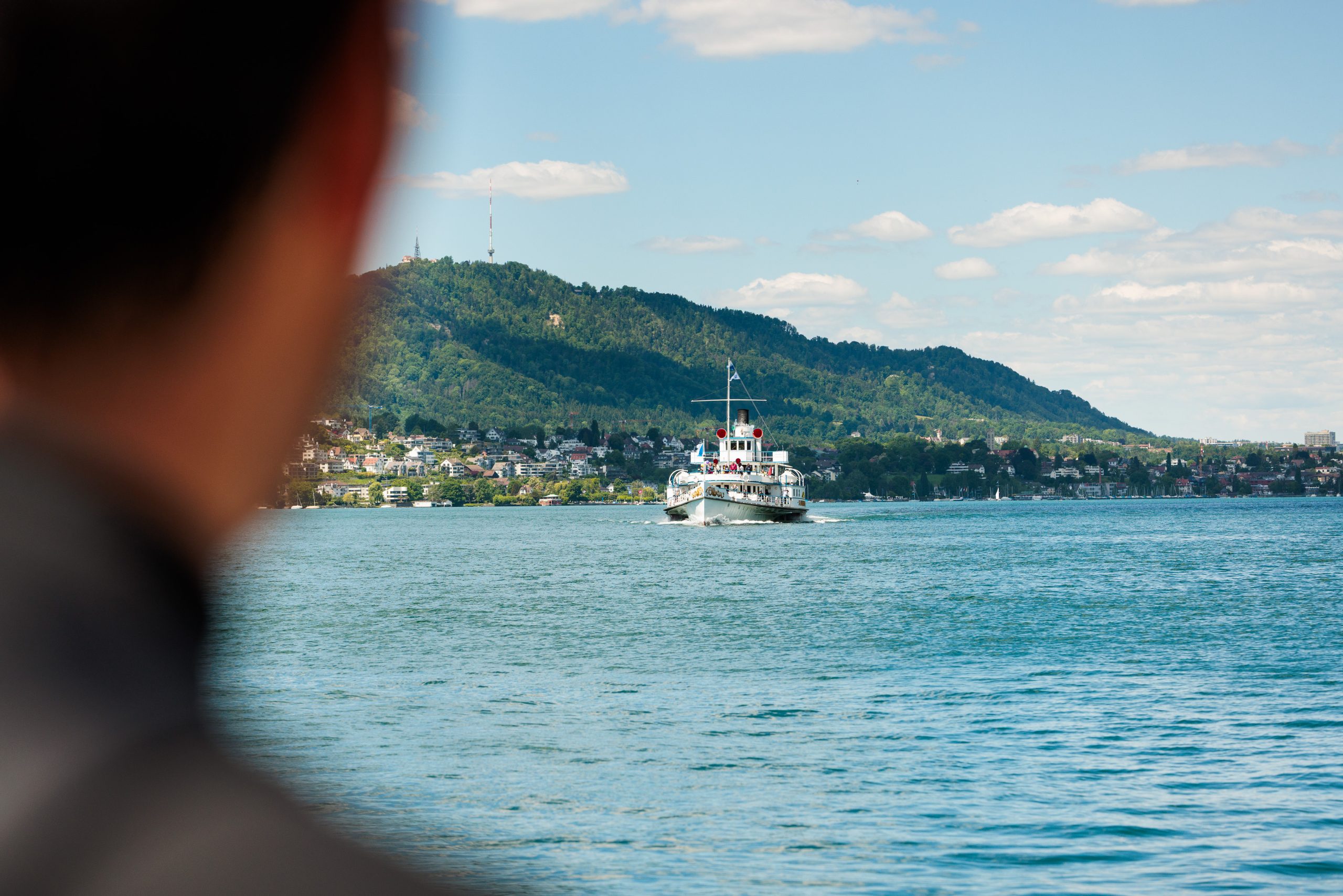 Eine Person im Vordergrund beobachtet ein weißes Fährboot auf einem See, mit grünen Hügeln und einer Stadt im Hintergrund unter einem teilweise bewölkten Himmel.