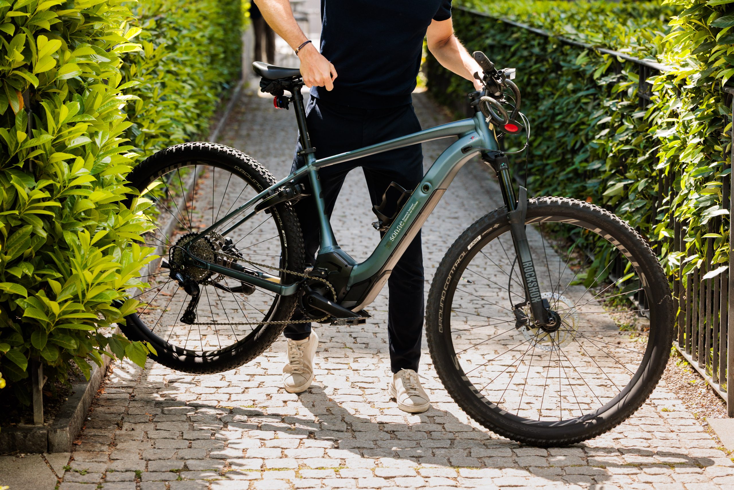 Eine dunkel gekleidete Person steht auf einem von grünen Hecken gesäumten Kopfsteinpflasterweg und hält ein modernes Mountainbike am Lenker aufrecht.