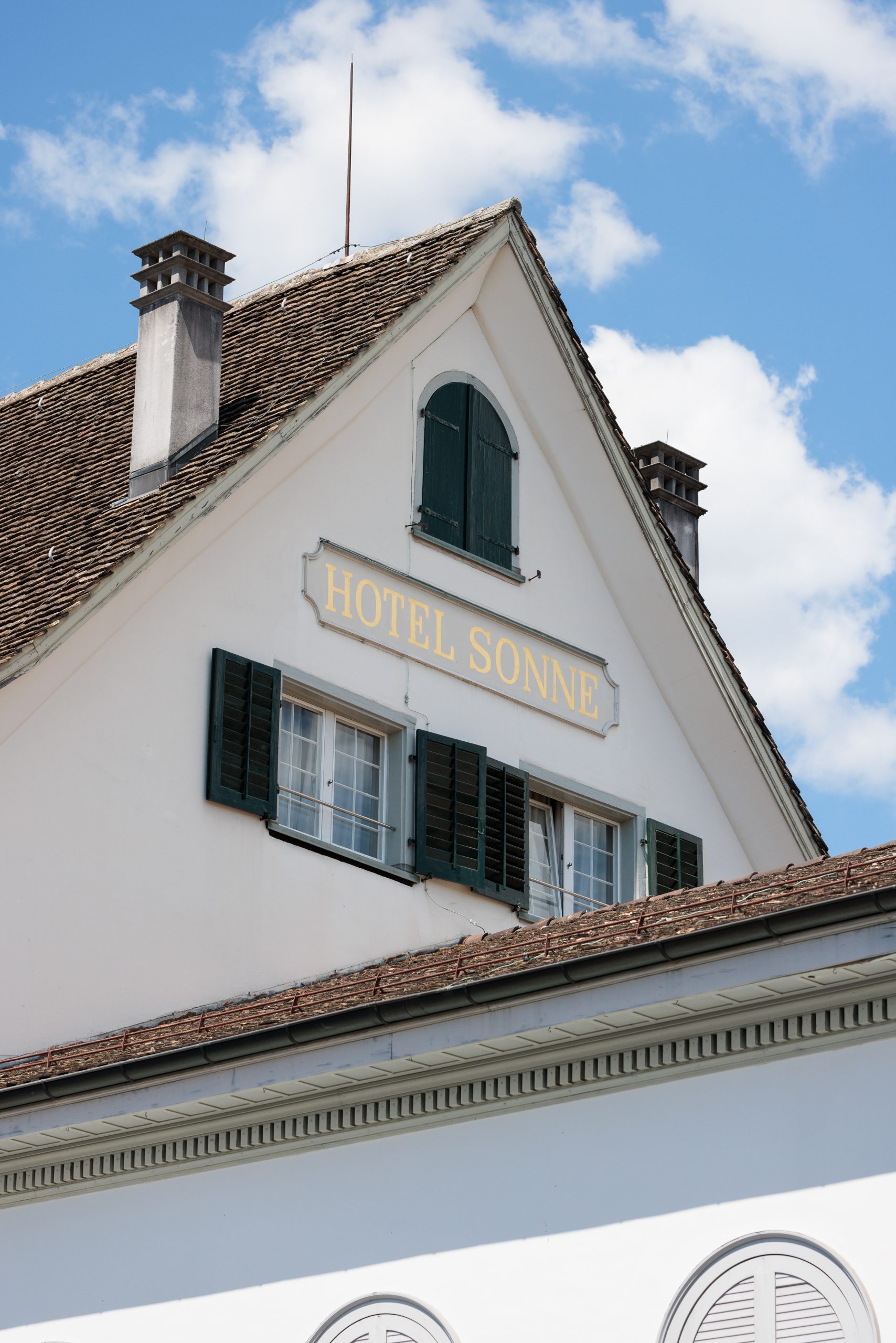 Ein weißes Gebäude mit dunkelgrünen Fensterläden und einem Schild mit der Aufschrift HOTEL SONNE unter dem Dach, mit blauem Himmel und vereinzelten Wolken im Hintergrund.