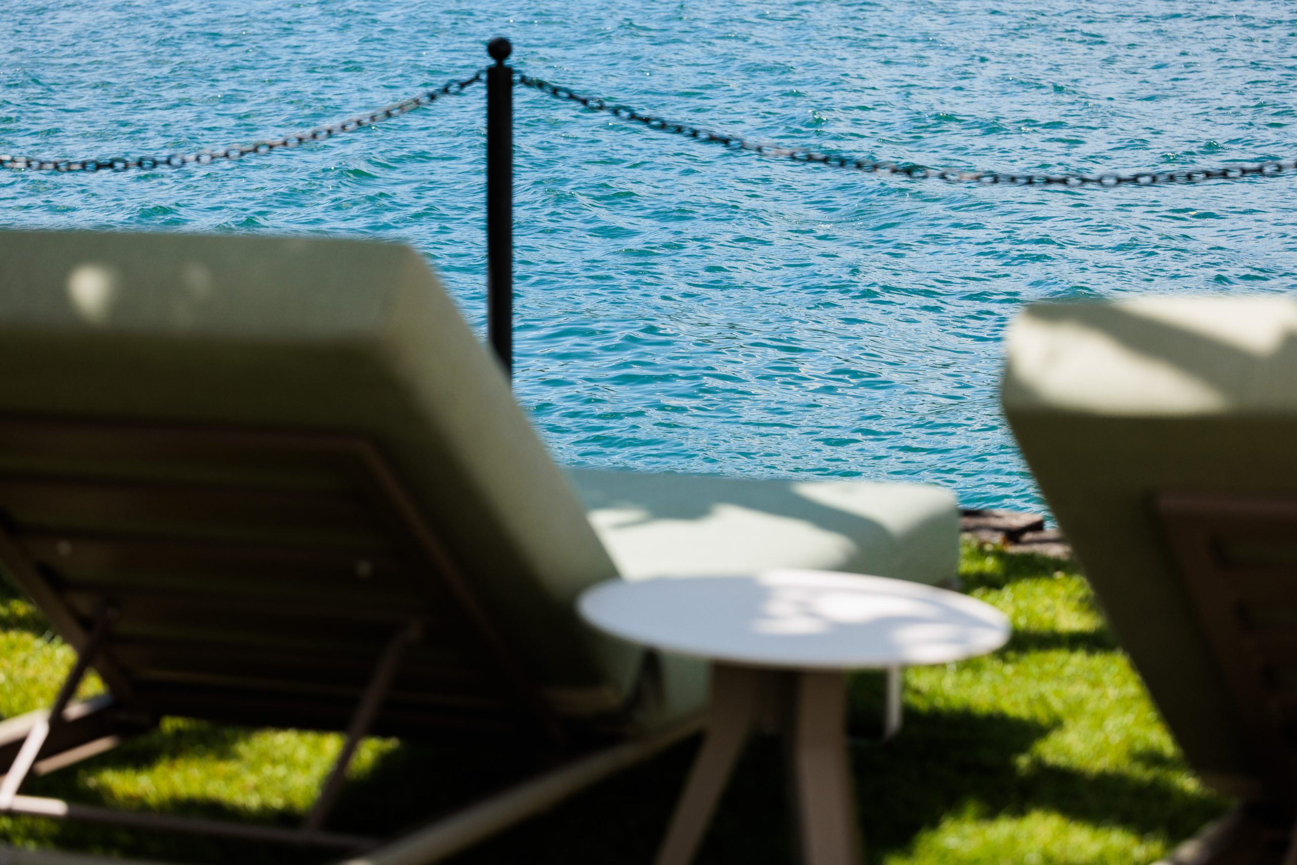 Zwei Liegestühle und ein kleiner Tisch im Gras stehen vor einem blauen Gewässer, das durch einen Kettenzaun getrennt ist. Die Szene suggeriert einen entspannenden Aufenthalt im Freien am Wasser.