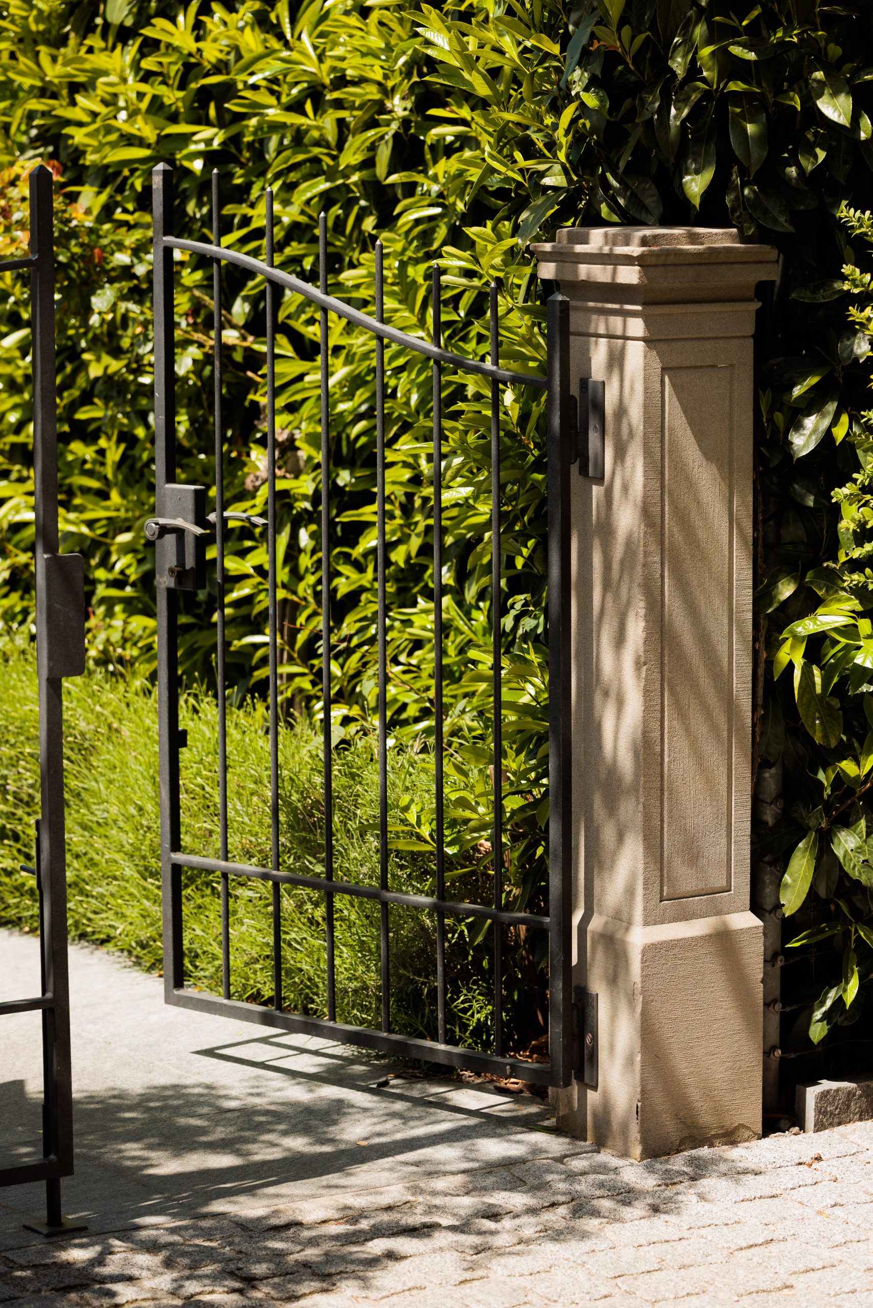Ein schwarzes Metalltor, das an einem beigen Steinpfeiler befestigt ist, steht neben einer üppigen grünen Hecke offen und wirft Schatten auf einen sonnenbeschienenen Steinweg.