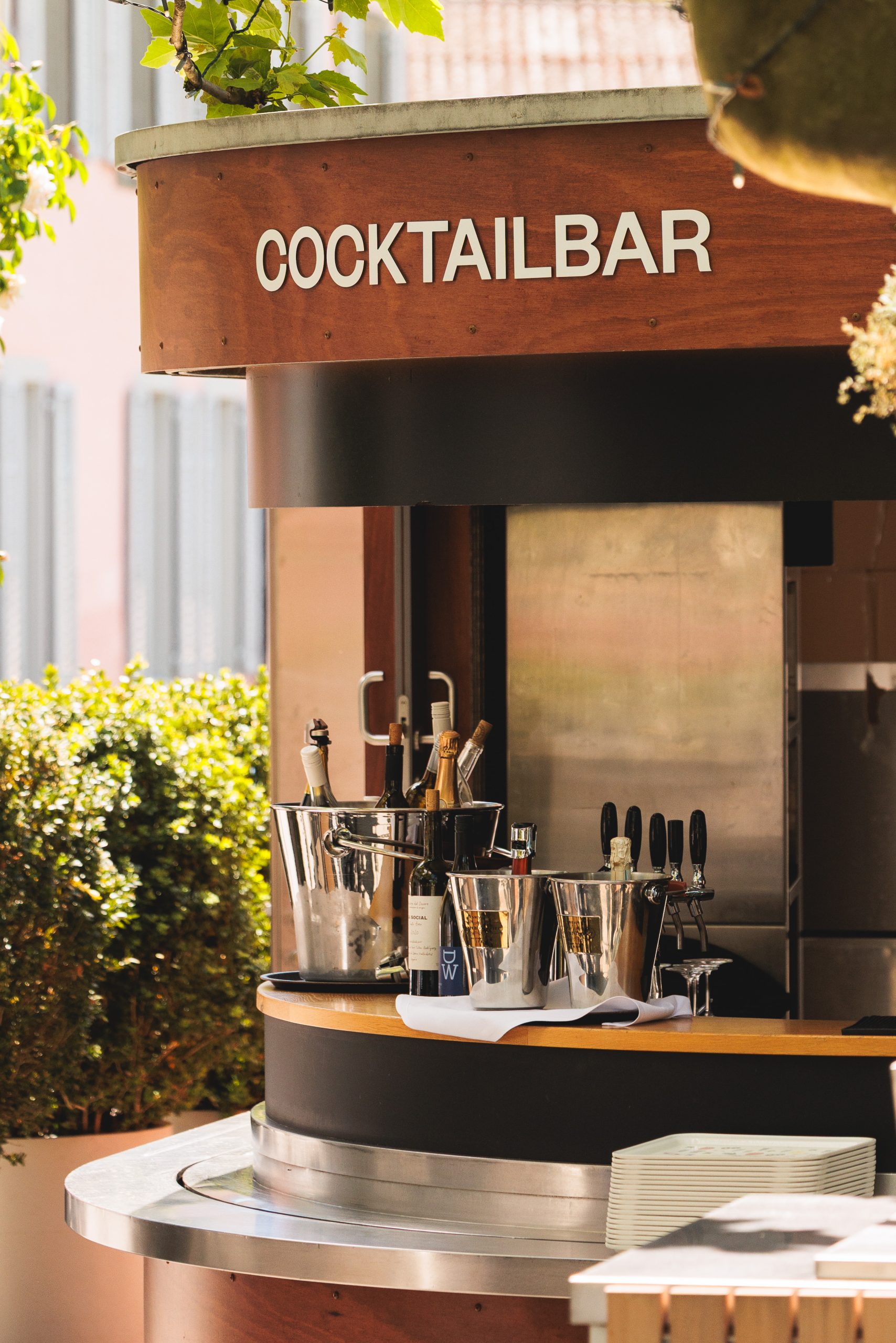 Eine stilvolle Cocktailbar im Freien mit einem geschwungenen Holztresen, Eiskübeln aus Metall, in denen Flaschen und Gläser stehen, und weißen Tellern, die an der Seite gestapelt sind, inmitten von Grünpflanzen. Auf dem Schild darüber steht COCKTAILBAR.