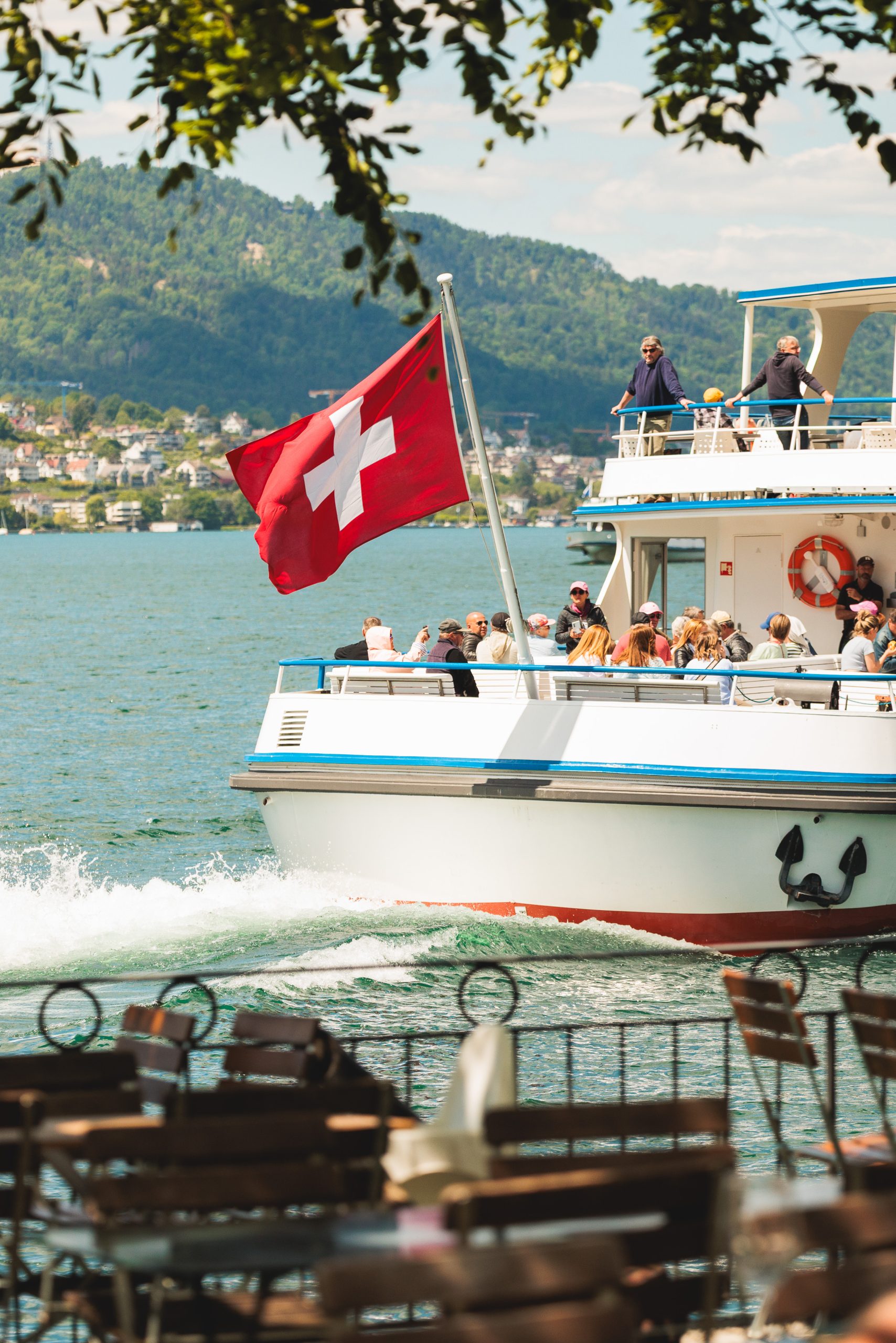 Ein Boot mit einer großen Schweizer Flagge fährt auf einem See und befördert Passagiere. Im Vordergrund ist das Ufer mit leeren Tischen und Stühlen zu sehen, während im Hintergrund grüne Hügel und eine Stadt zu sehen sind.