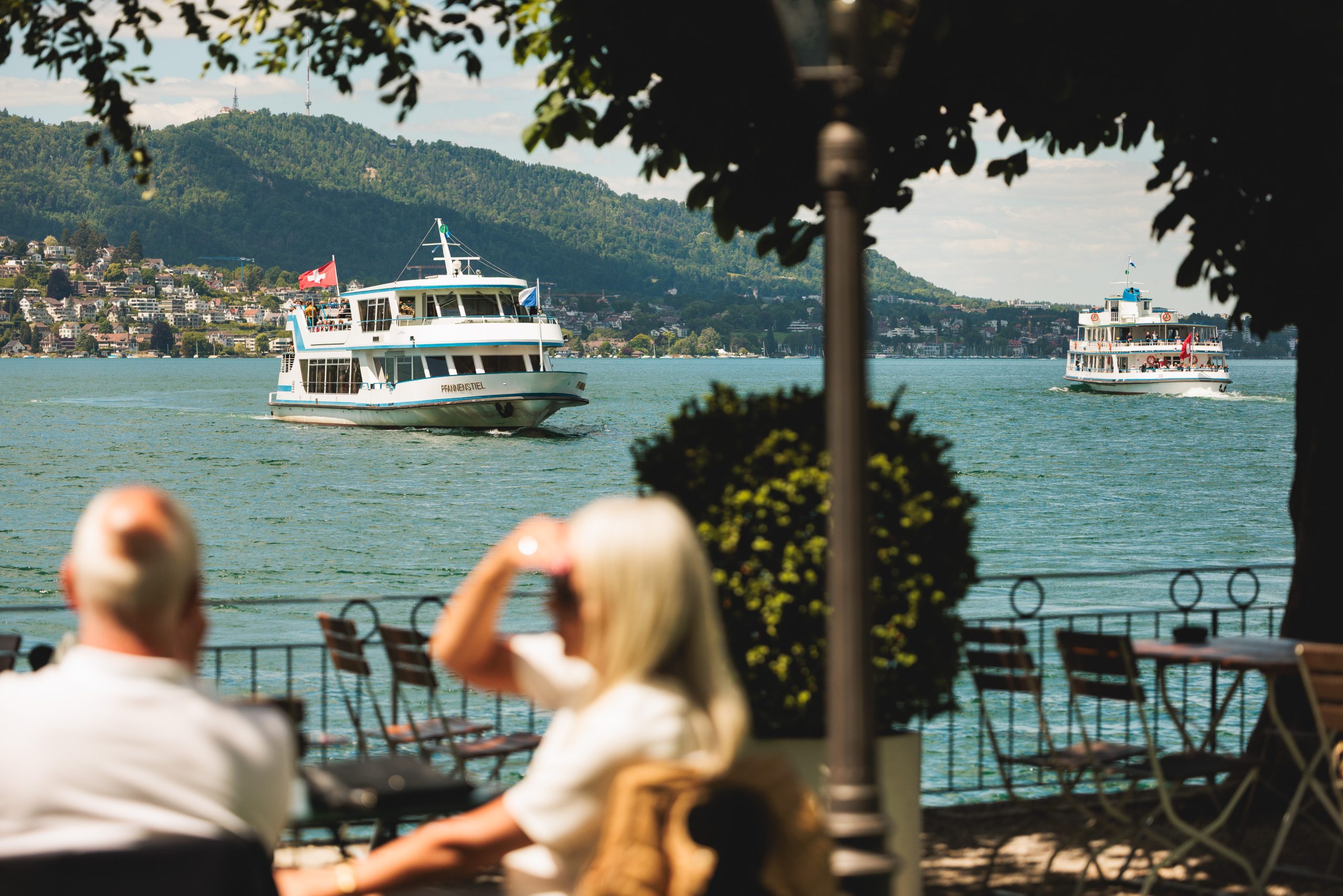 Zwei Personen sitzen in einem Straßencafé an einem See, auf dem zwei Boote fahren. Im Hintergrund sind unter einem teilweise bewölkten Himmel Hügel und Häuser zu sehen. Bäume und Pflanzen rahmen die Szene ein.