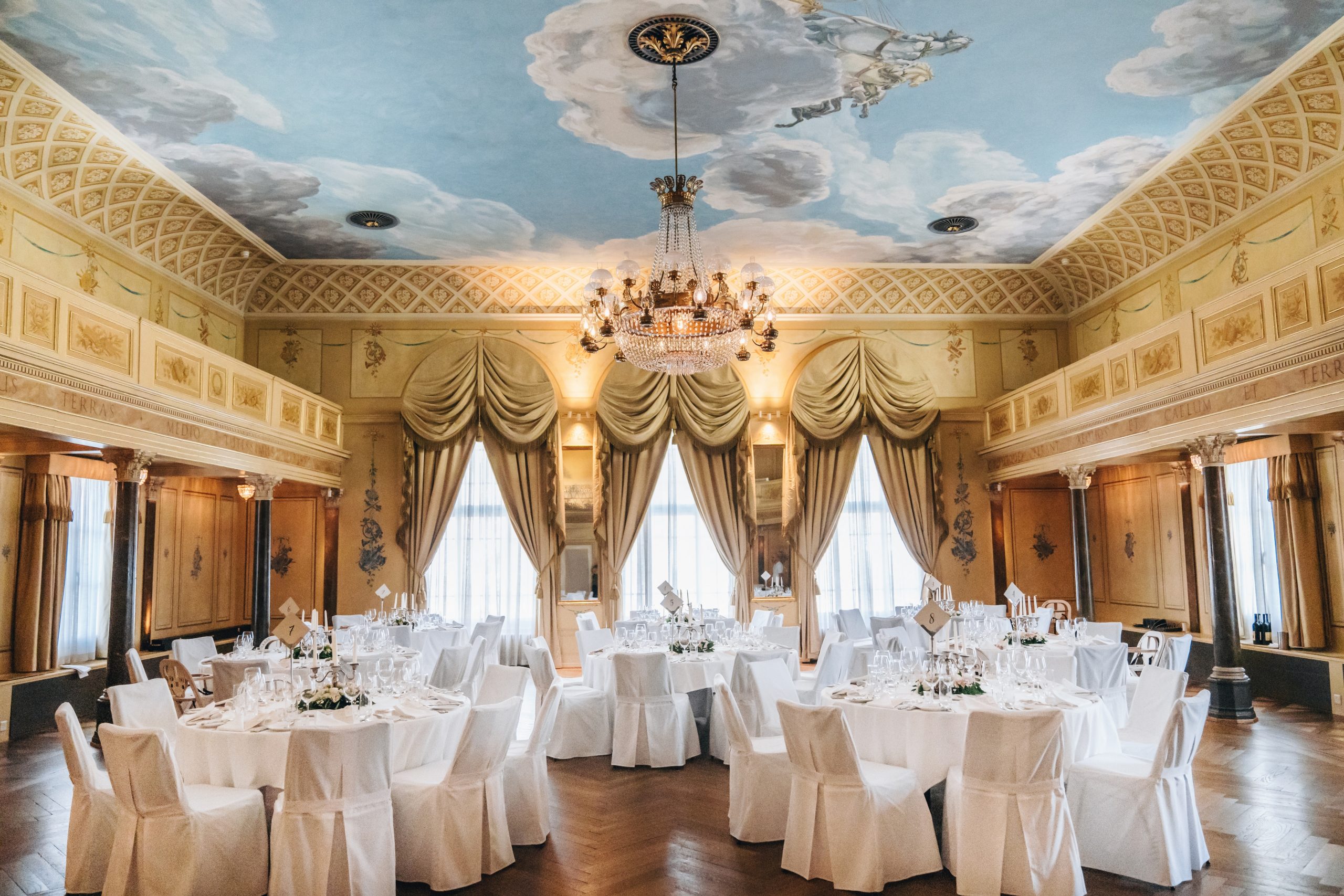 Ein eleganter Bankettsaal mit runden, weiß gedeckten Tischen, weißen Stühlen, großen Fenstern mit goldenen Vorhängen, einem Kronleuchter, verschnörkelten gelben Wänden und einer mit Wolken und einer Putte bemalten Decke.