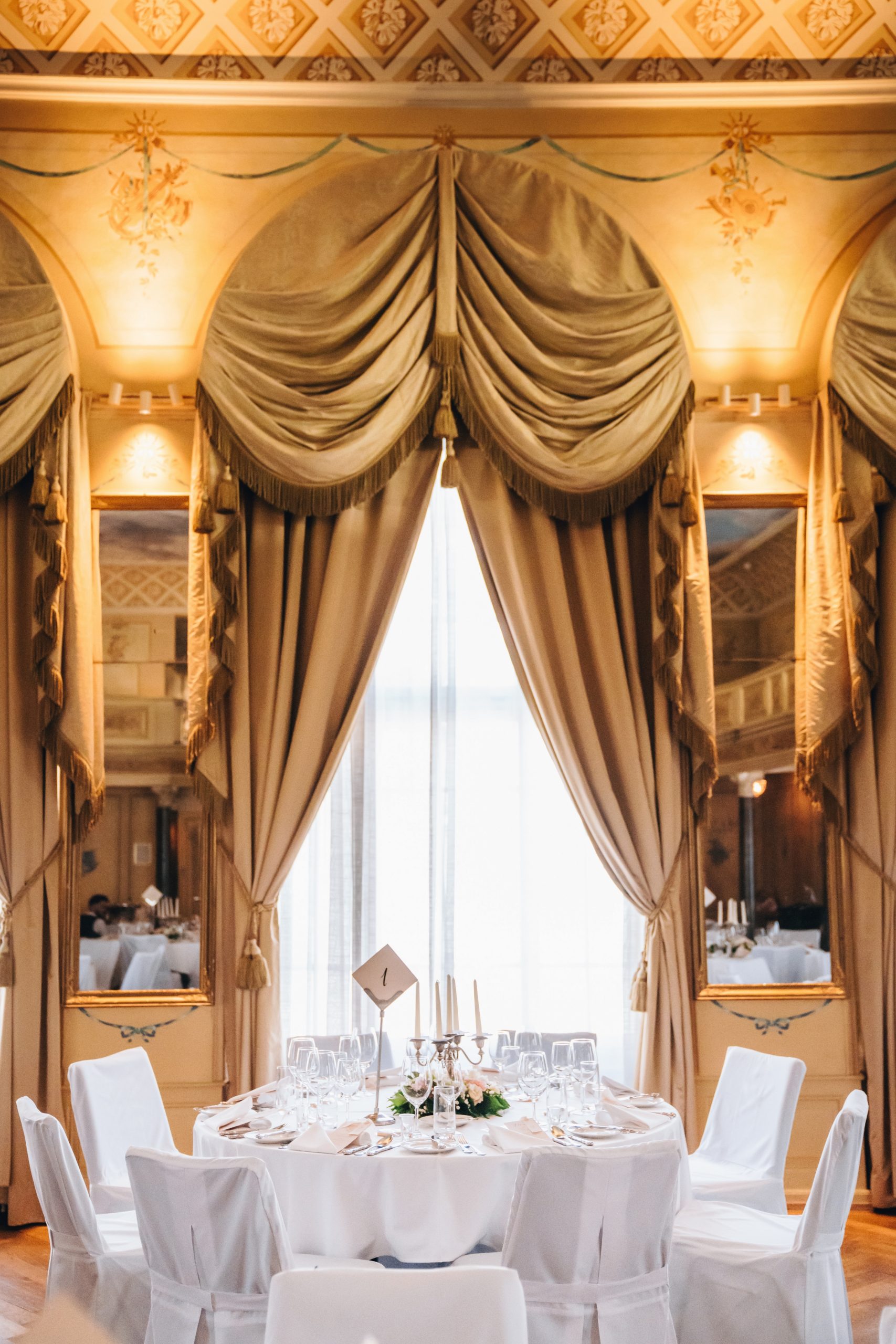 Ein runder Tisch, gedeckt für eine formelle Veranstaltung mit weißen Stühlen, elegantem Geschirr und einem blumengeschmückten Tafelaufsatz, platziert vor großen verzierten goldenen Vorhängen und Spiegeln in einem prunkvollen, klassischen Raum.